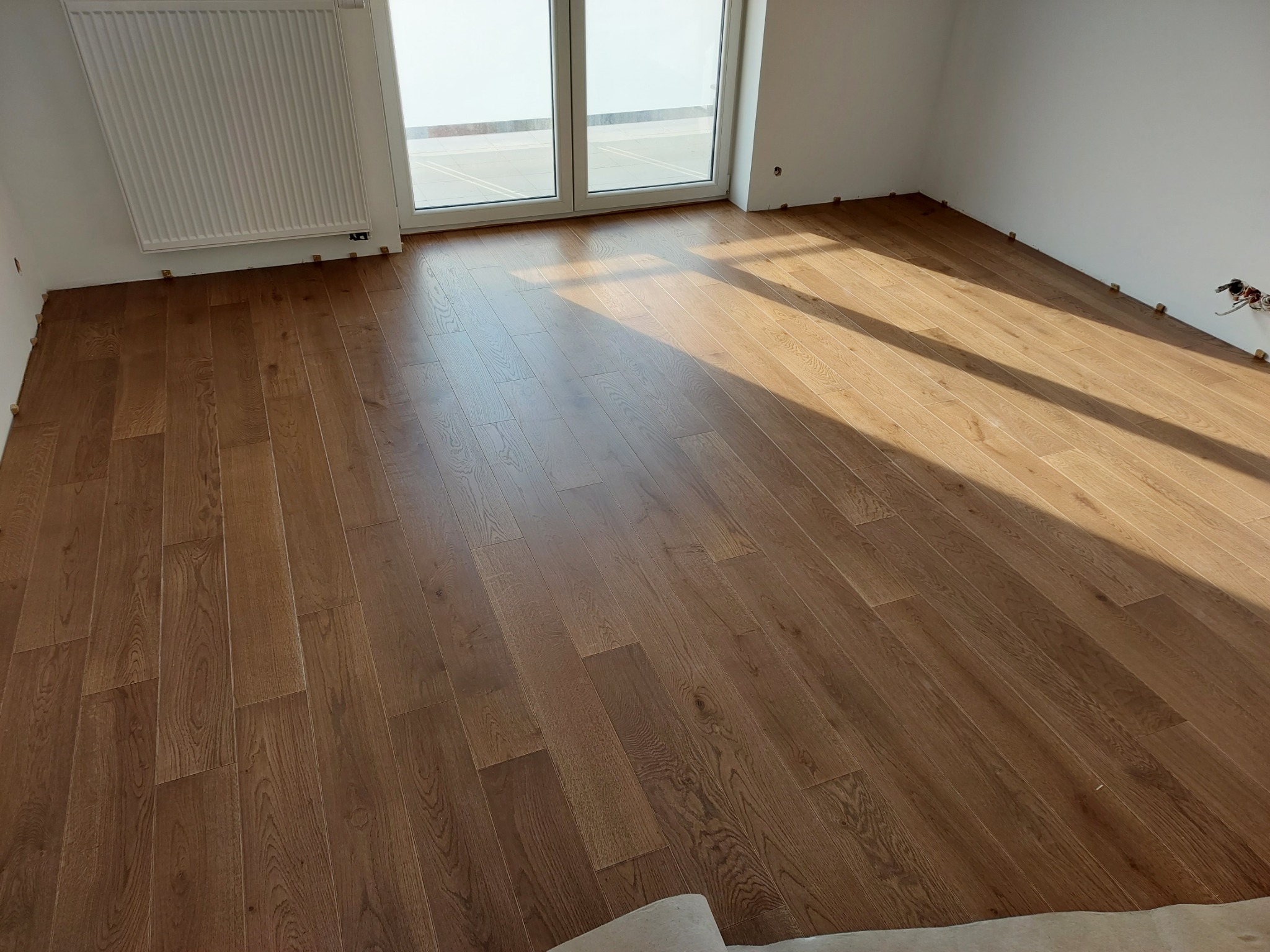 Ułożony parkiet w jasnym odcieniu drewna w nowym pokoju, widoczne listwy przypodłogowe czekające na montaż, zarys okna balkonowego w tle.