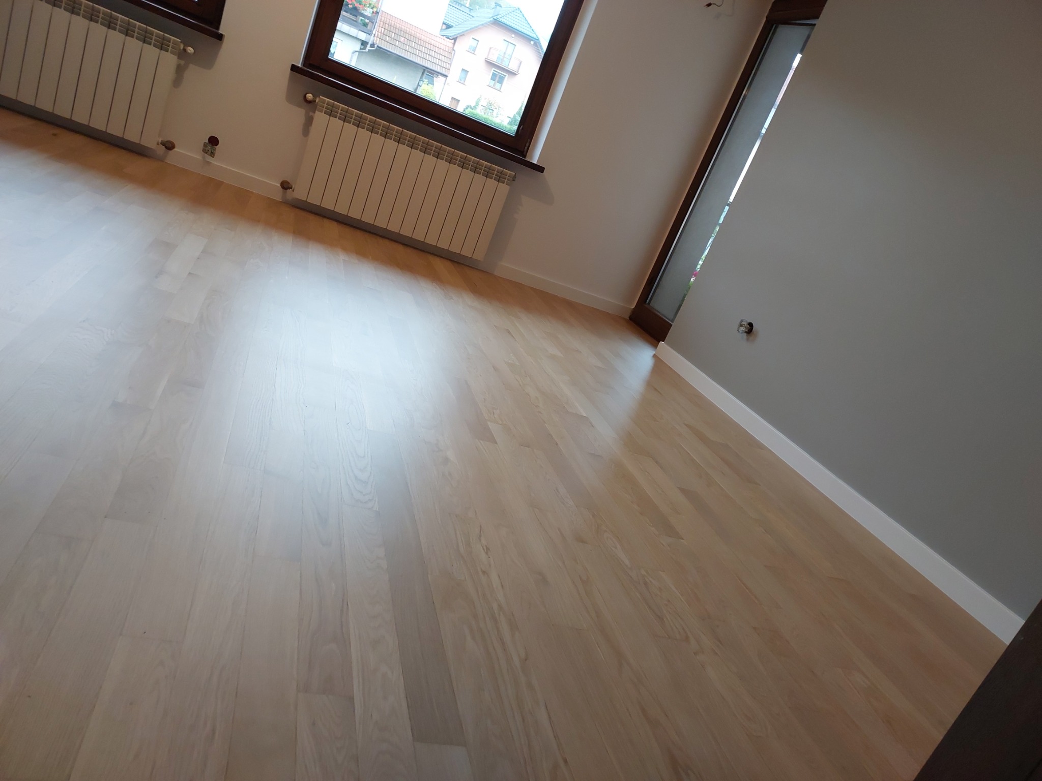 Jasny, świeżo ułożony parkiet w nowym mieszkaniu, widok z ukosa, grzejniki pod oknami i szara ściana w tle.