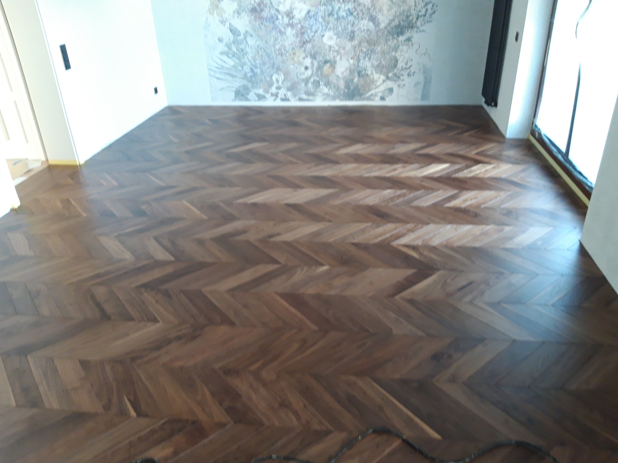 Ułożony parkiet w jodełkę z ciemnego drewna, widoczna ściana z delikatnym wzorem kwiatowym i listwa przypodłogowa.