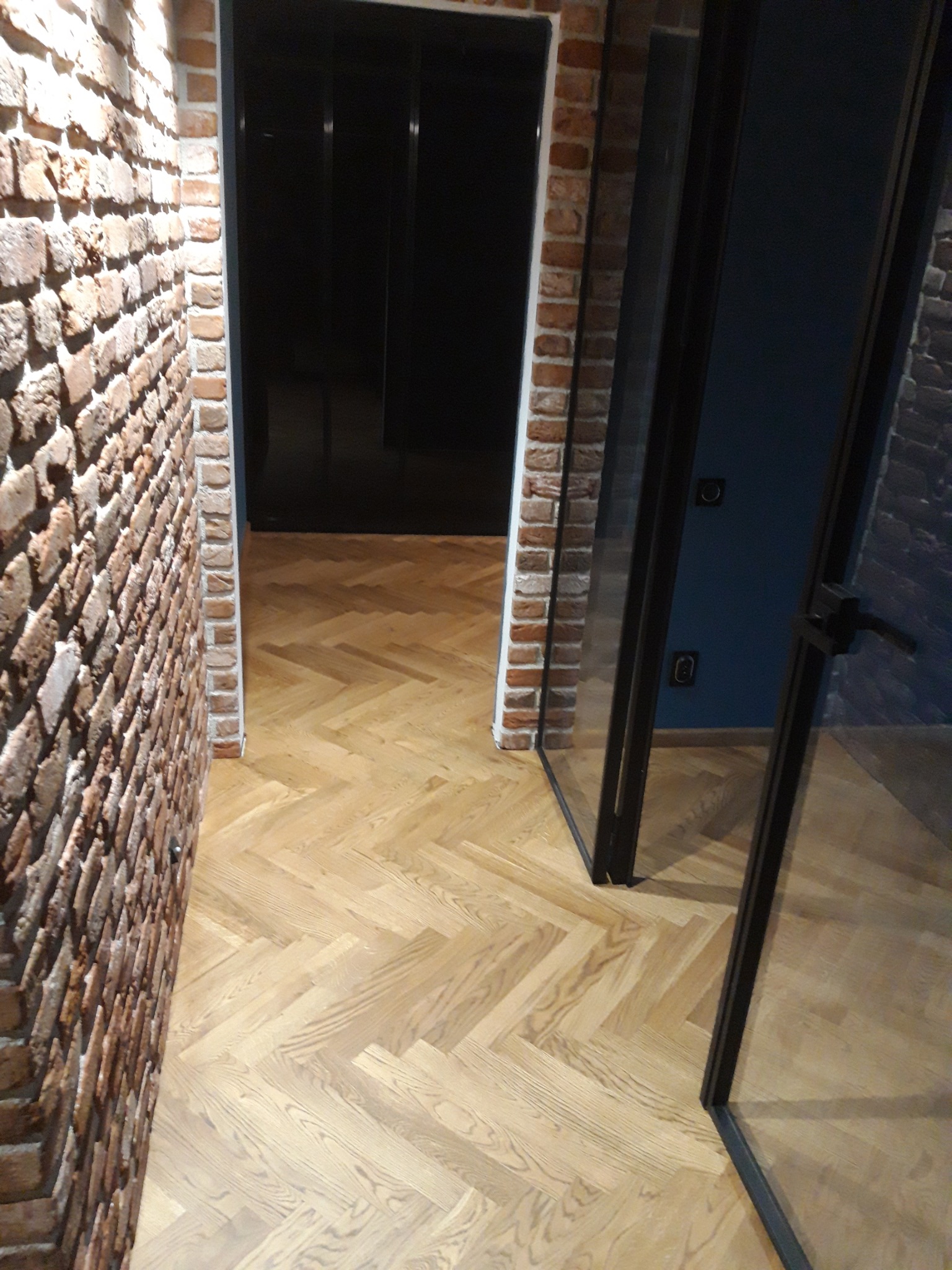 Ułożony parkiet w jodełkę, jasne drewno, widok z perspektywy korytarza z ceglanymi ścianami i szklanymi drzwiami z czarnymi ramami.