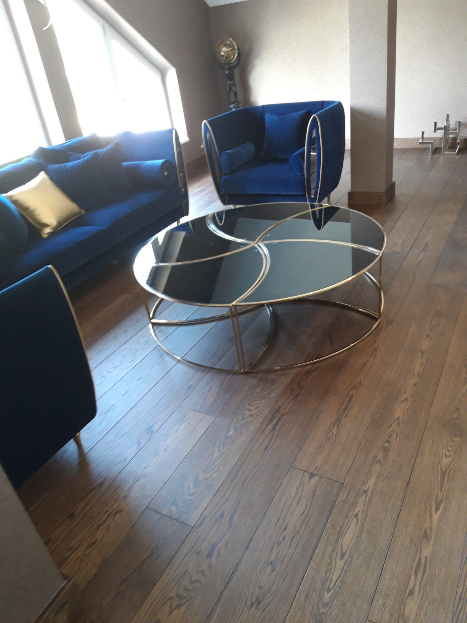 Salon z ciemną, drewnianą podłogą z parkietu ułożonego w deski, na której stoi okrągły, szklany stolik kawowy ze złotymi nogami. W tle niebieski komplet wypoczynkowy z sofą i fotelem.