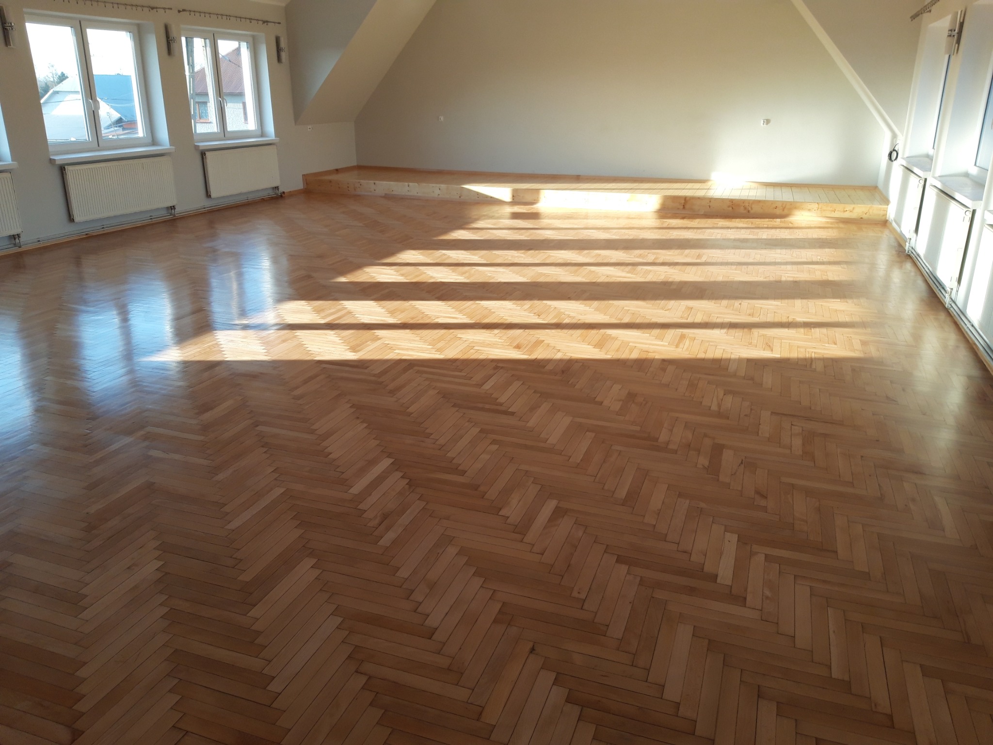 Jasny, przestronny pokój na poddaszu z parkietem ułożonym we wzór jodełki francuskiej, zalany popołudniowym światłem słonecznym wpadającym przez okna, tworzącym wyraźne cienie.