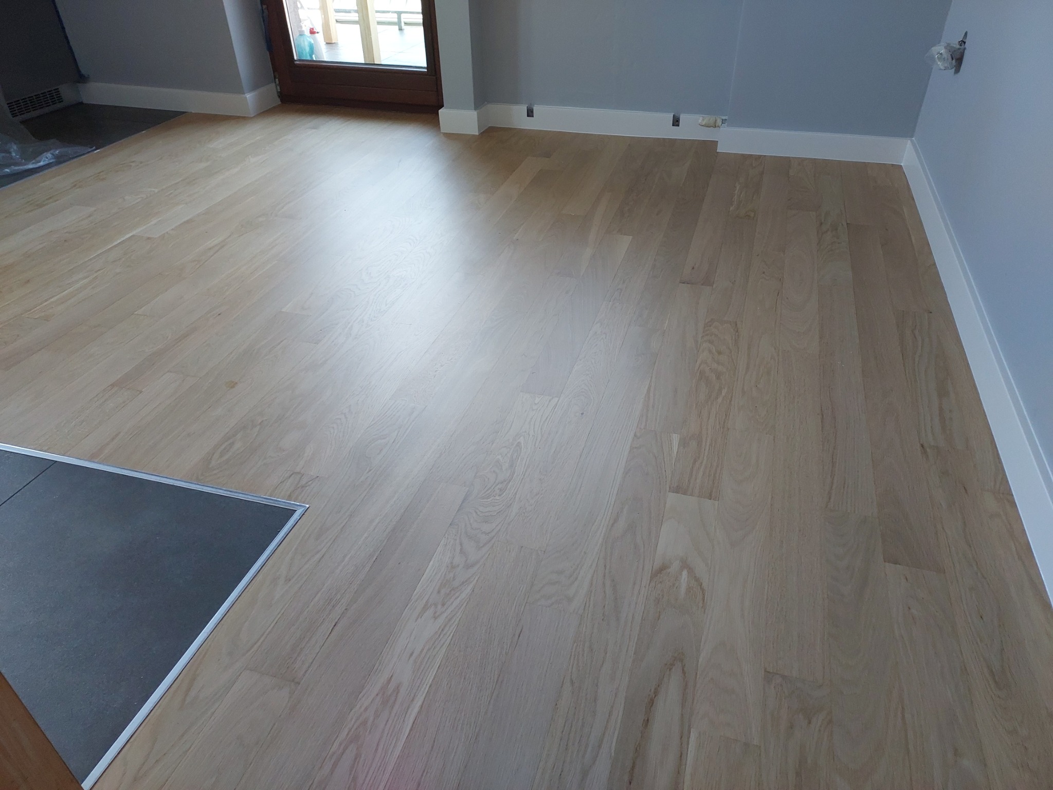 Świeżo ułożony, jasny parkiet w pokoju, widoczny fragment ciemnoszarej podłogi przy wejściu, białe listwy przypodłogowe i szare ściany.