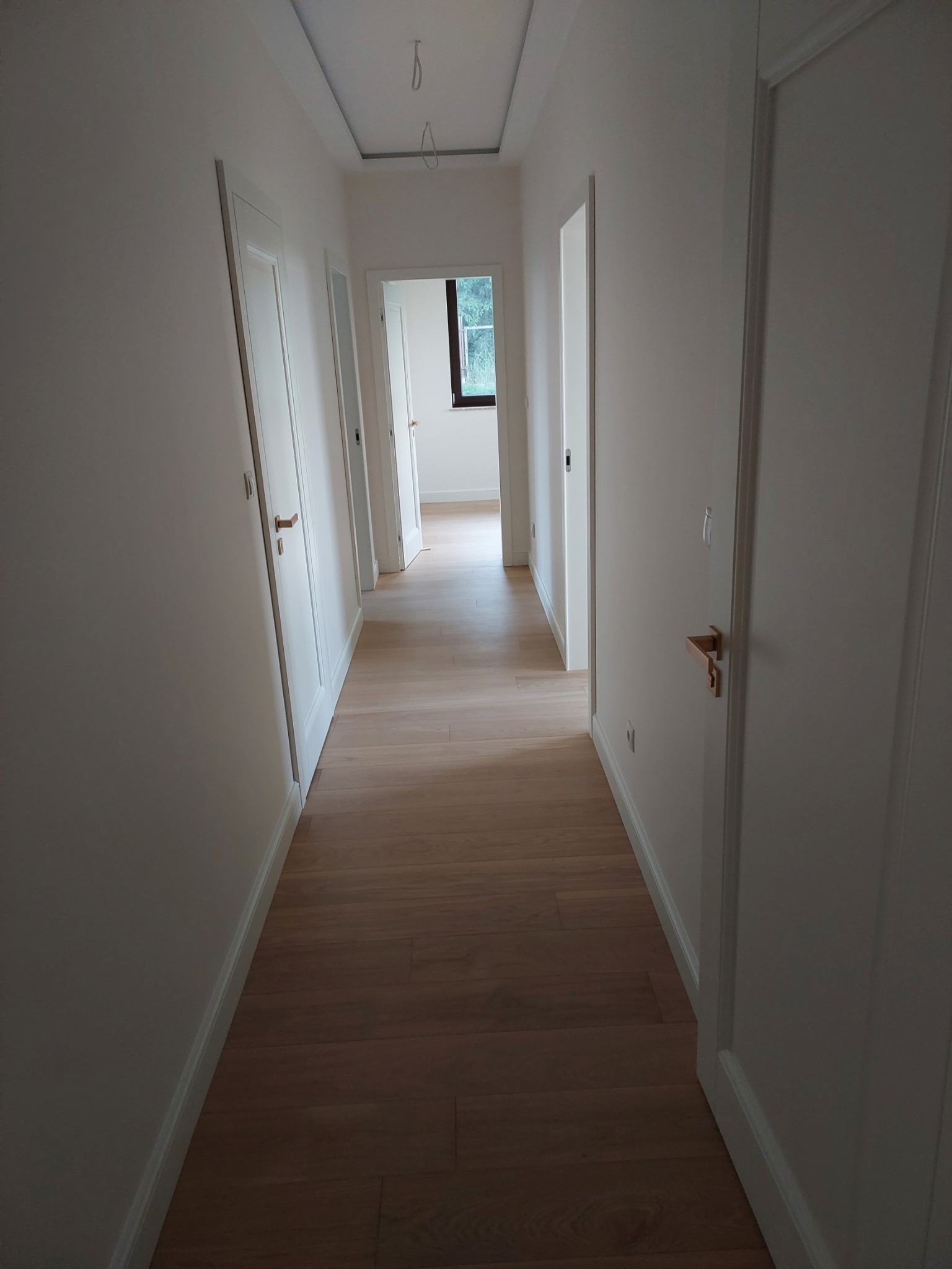 Długi korytarz z jasnym parkietem ułożonym w deski, białymi ścianami i białymi drzwiami z minimalistycznymi, drewnianymi klamkami, widok w głąb z oknem na końcu.