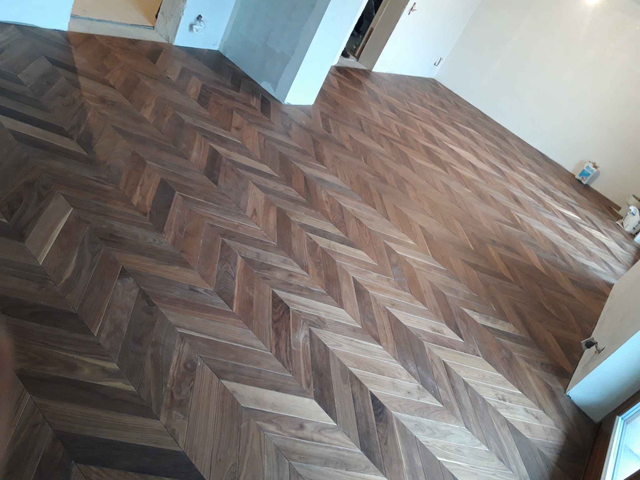 Ułożony parkiet w jodełkę z ciemnego drewna, widoczne niedoskonałości na łączeniach i drobne zabrudzenia, ściany w trakcie wykończenia.