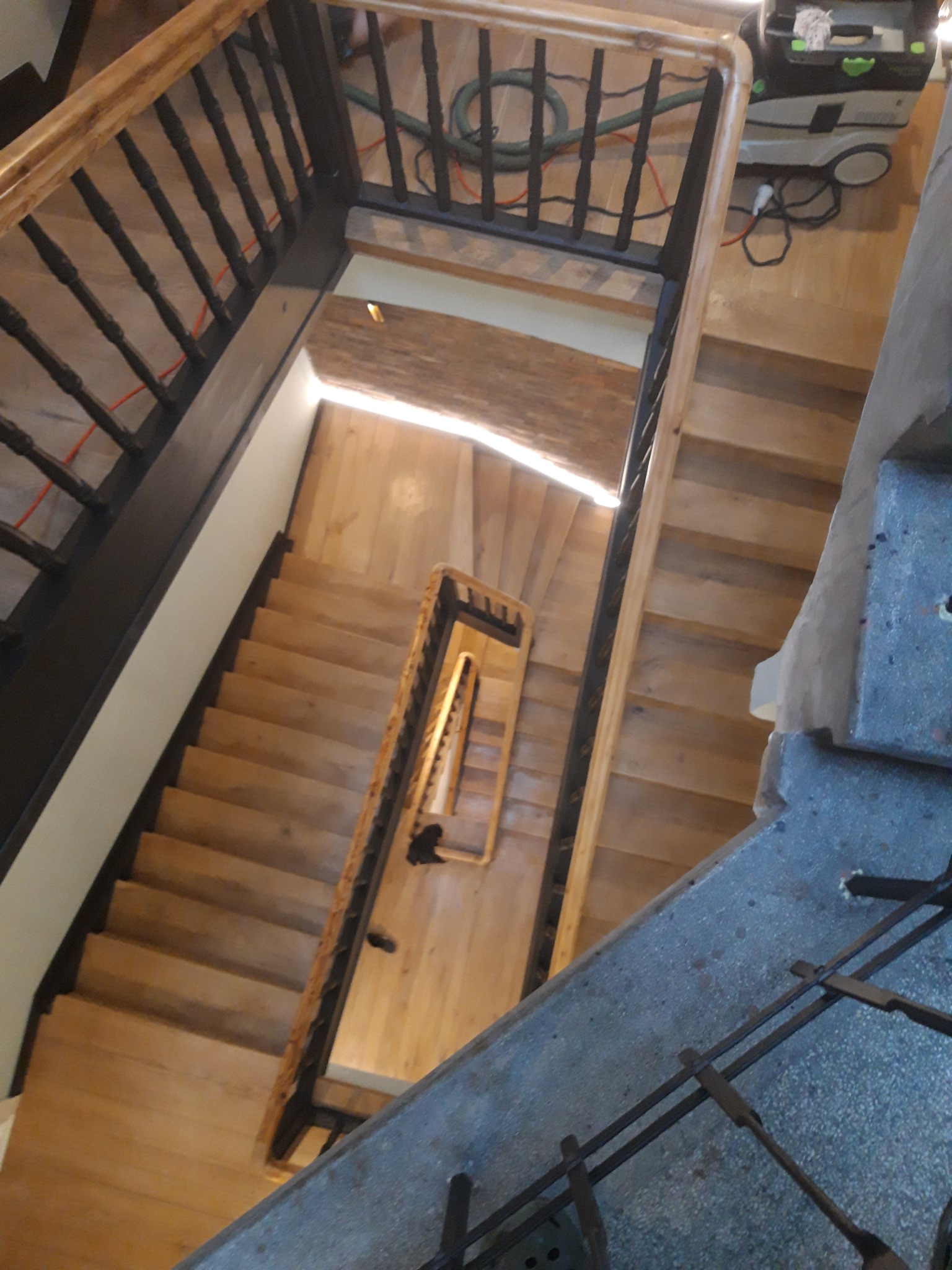 Widok z góry na drewniane schody podczas renowacji, z widocznym odkurzaczem przemysłowym i przewodem na podłodze, sugerujący prace związane z czyszczeniem lub przygotowaniem powierzchni.