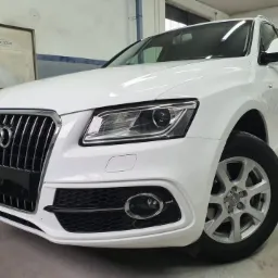 Biały samochód Audi Q5, widok z przodu, z widocznymi detalami karoserii i felgami, w warsztacie z metalową szafką w tle.