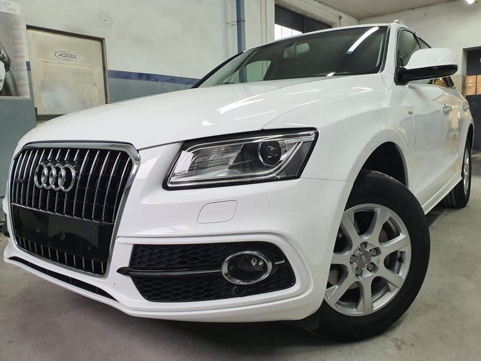 Biały samochód Audi Q5, widok z przodu, z widocznymi detalami karoserii i felgami, w warsztacie z metalową szafką w tle.