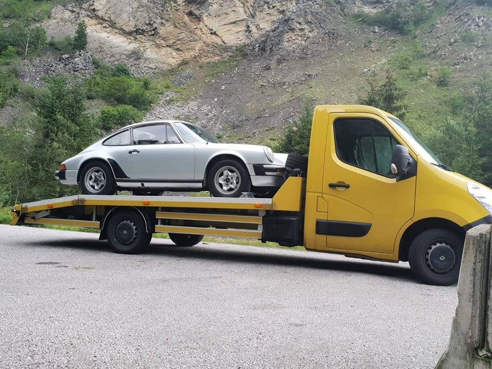 Żółta laweta przewozi srebrne Porsche 911 na tle skalistego zbocza porośniętego zielenią.