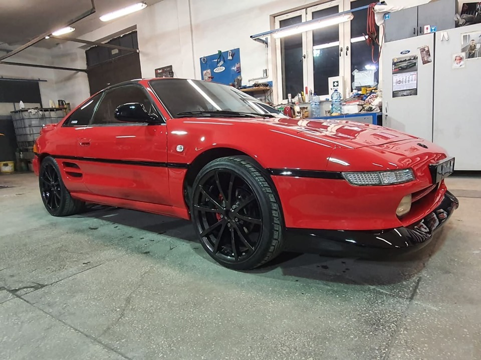 Czerwone sportowe auto z czarnymi felgami, stojące w warsztacie, widoczne detale karoserii i oświetlenie warsztatowe.