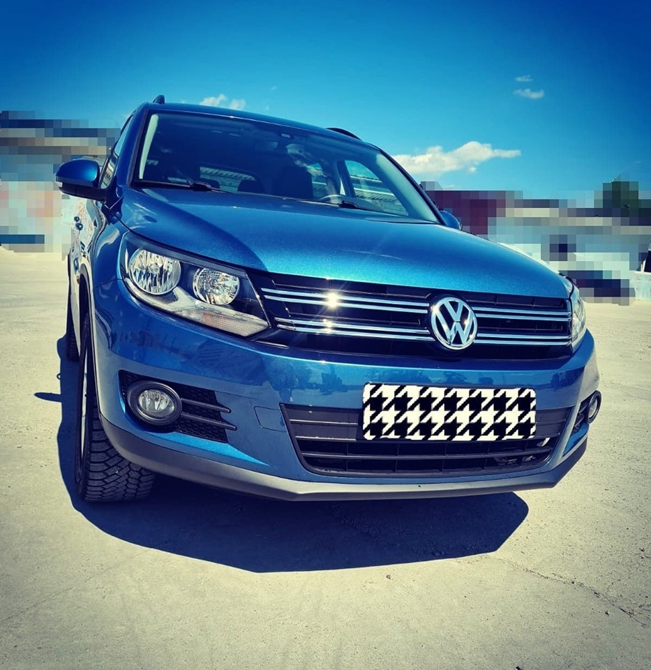 Błękitny Volkswagen Tiguan zaparkowany na betonowym placu w słoneczny dzień, z widoczną kratką na miejscu tablicy rejestracyjnej i delikatnym filtrem na zdjęciu.