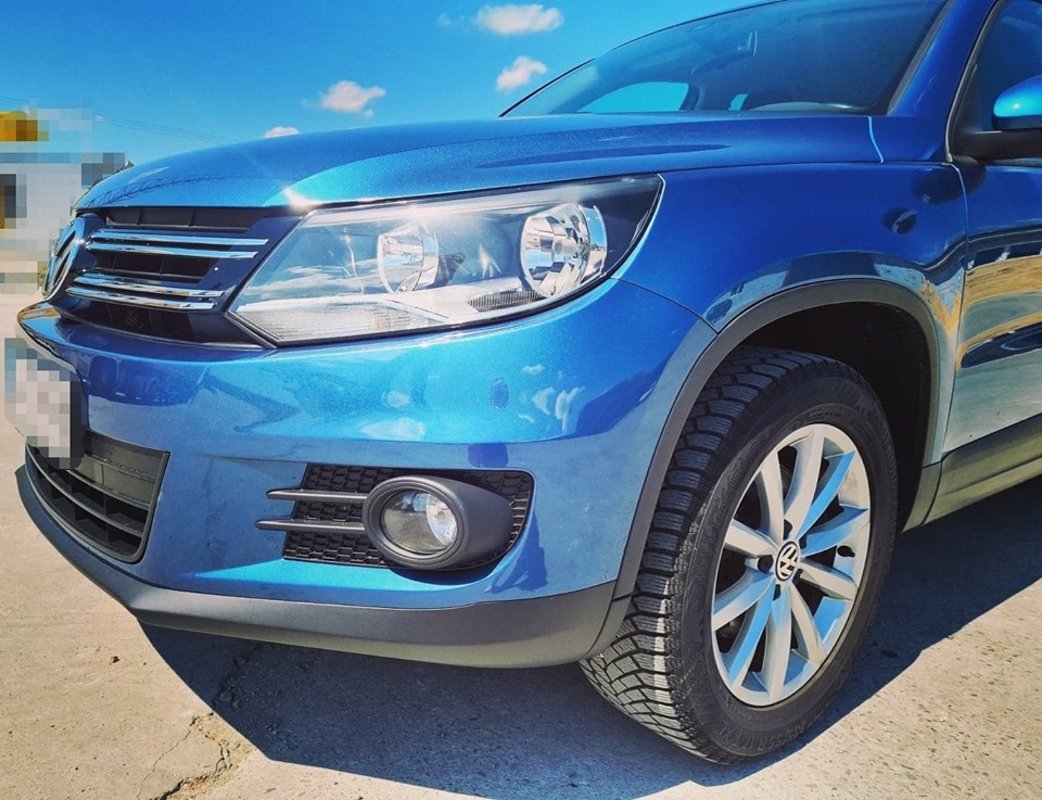 Błękitny samochód osobowy Volkswagen Tiguan, widok z przodu, z oponami zimowymi na betonowym podłożu w słoneczny dzień.