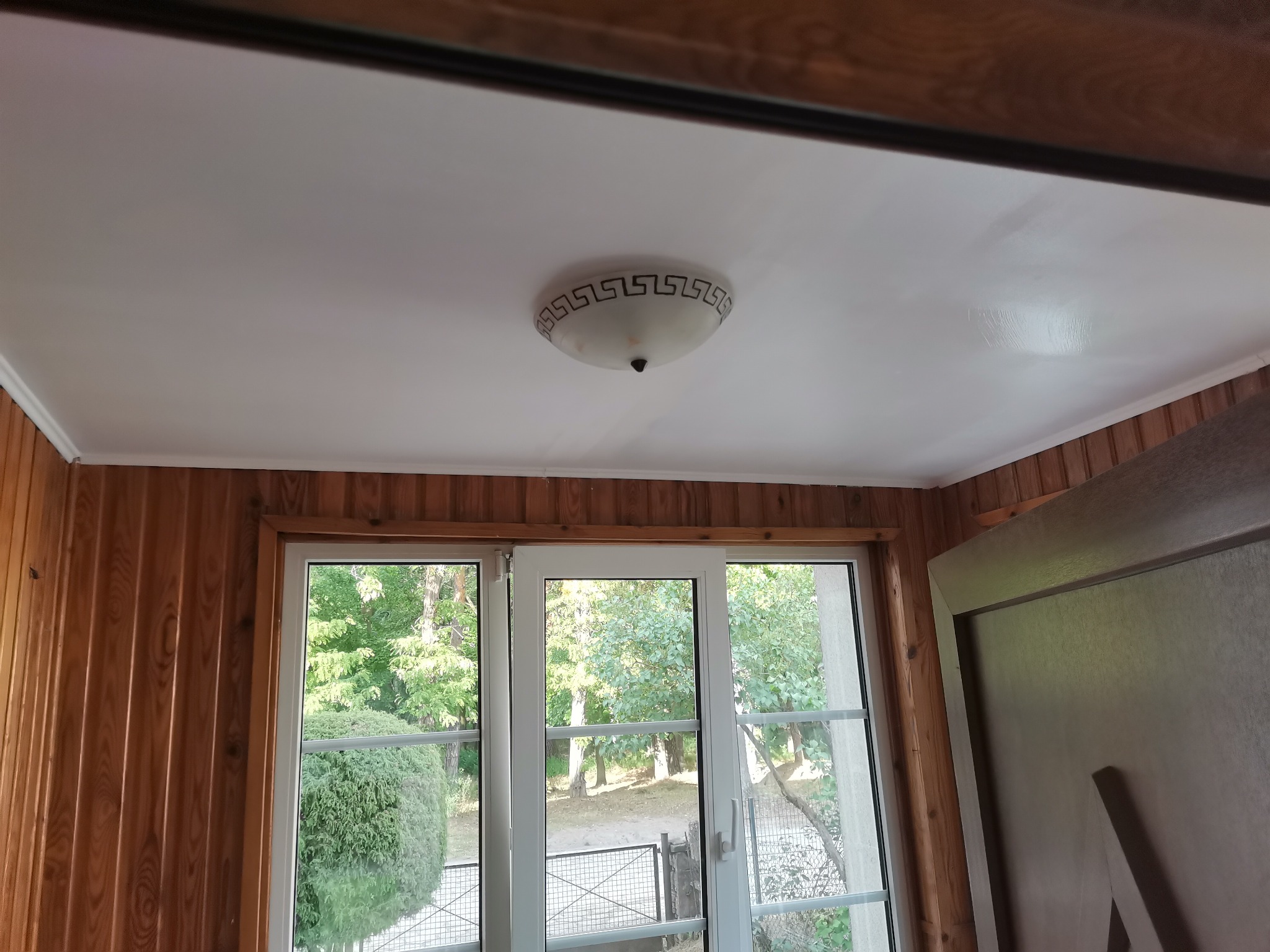 Biały sufit po malowaniu z widocznym śladem po wałku, lampa sufitowa z greckim wzorem, boazeria drewniana i okno z widokiem na zieleń.