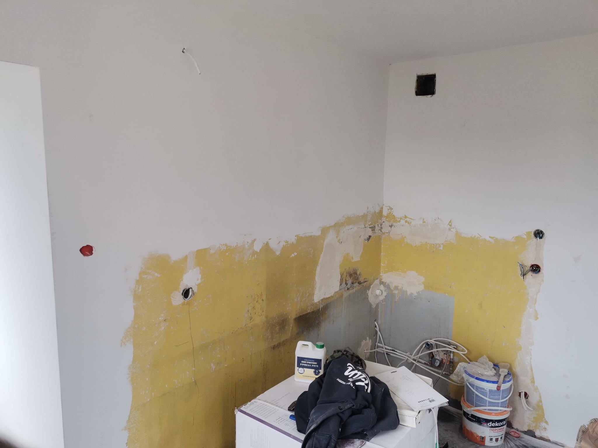 Ściana w trakcie przygotowania do malowania: widoczne fragmenty starej żółtej farby, gładź szpachlowa, puszki z farbą Dekoral i wiązka kabli elektrycznych wystająca ze ściany.