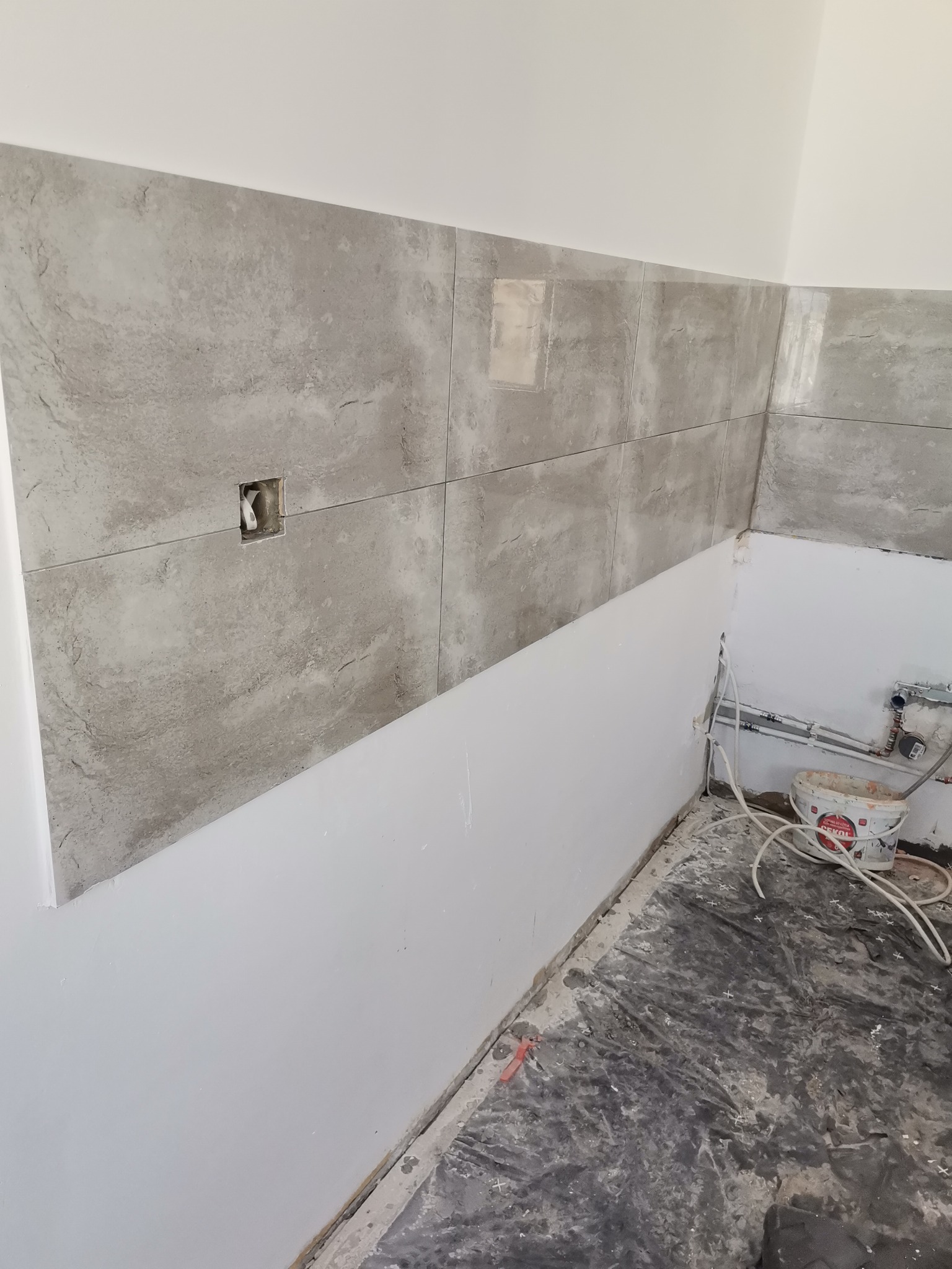 Układanie szarych płytek imitujących kamień na ścianie w trakcie prac w Gorzowie Wielkopolskim, widoczne puszki elektryczne i niedomalowana dolna część ściany, na podłodze folia ochronna.