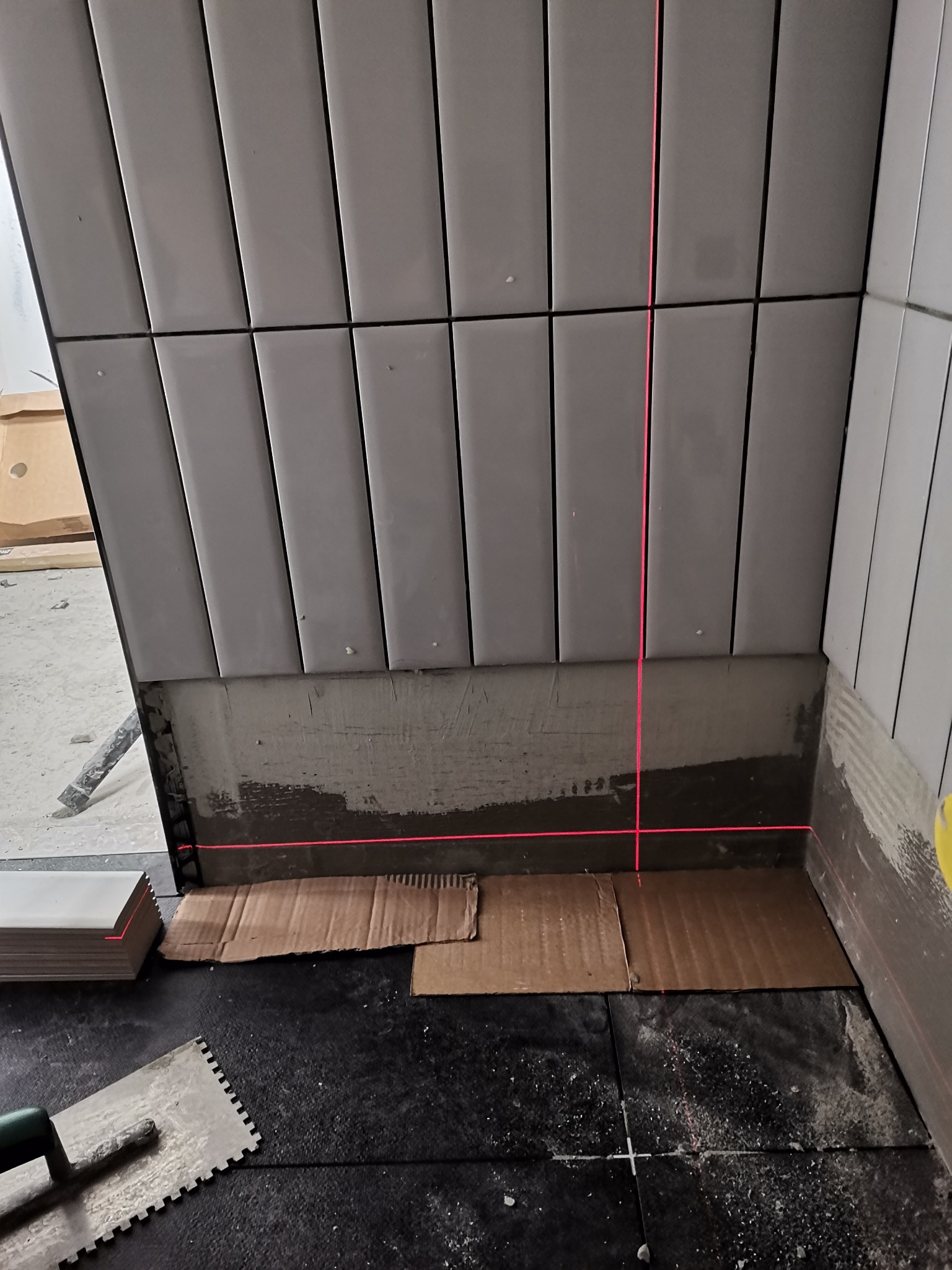 Układanie białych, prostokątnych płytek na ścianie w łazience, widoczny laser krzyżowy wyznaczający poziom, paczka płytek i kielnia na podłodze zabezpieczonej kartonem.