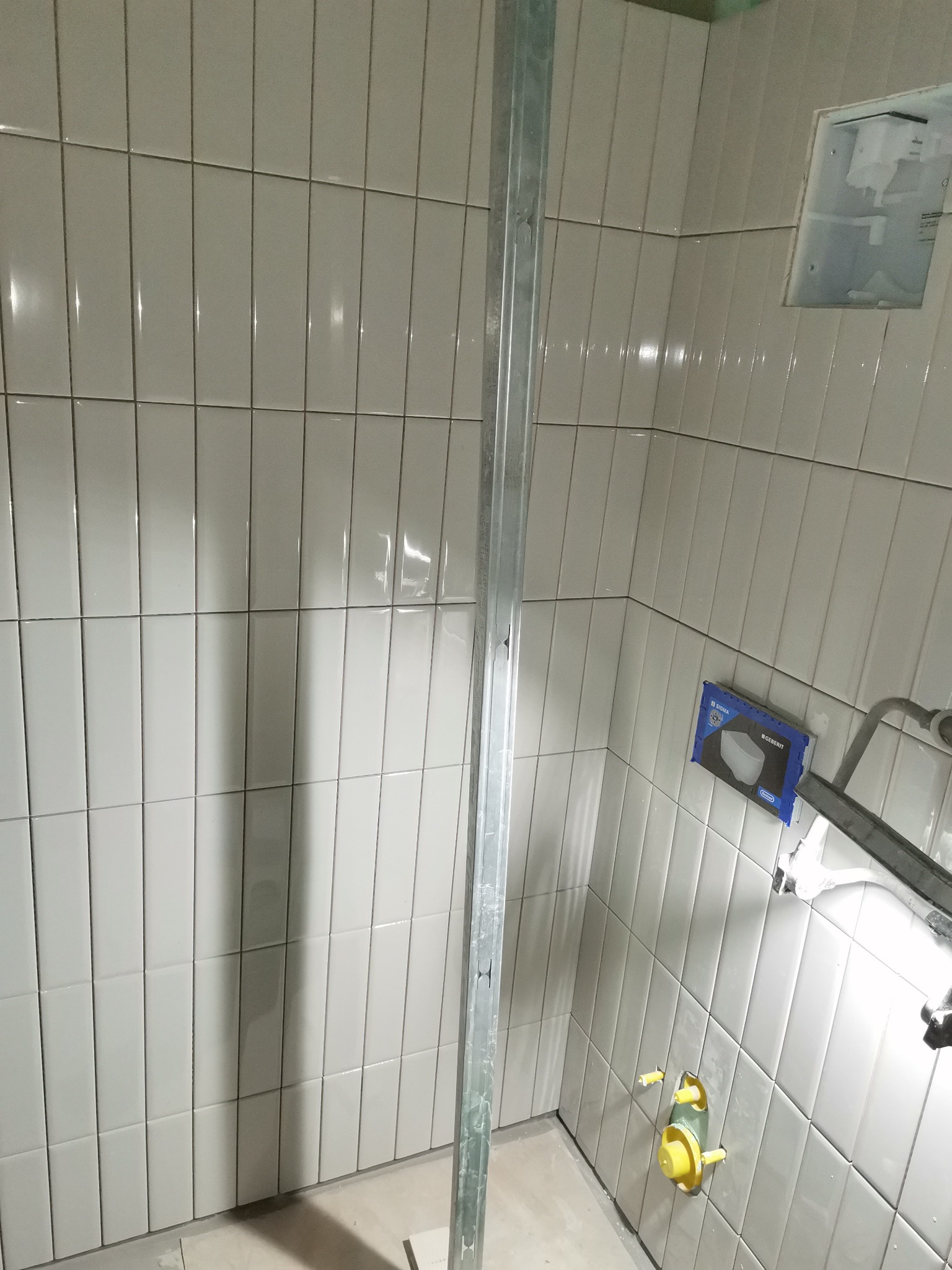 Ściany łazienki wyłożone pionowymi, białymi płytkami w trakcie remontu, widoczny metalowy profil, puszka podtynkową i elementy instalacji wodnej.