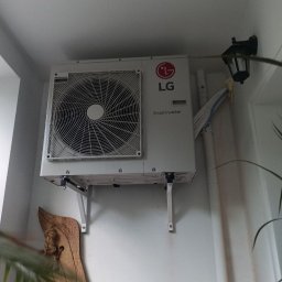 HVAC serwis - Biała jednostka zewnętrzna klimatyzacji LG Smart Inverter zamontowana na ścianie, widoczne kable i lampa ścienna. Dekoracja z drewna w tle.