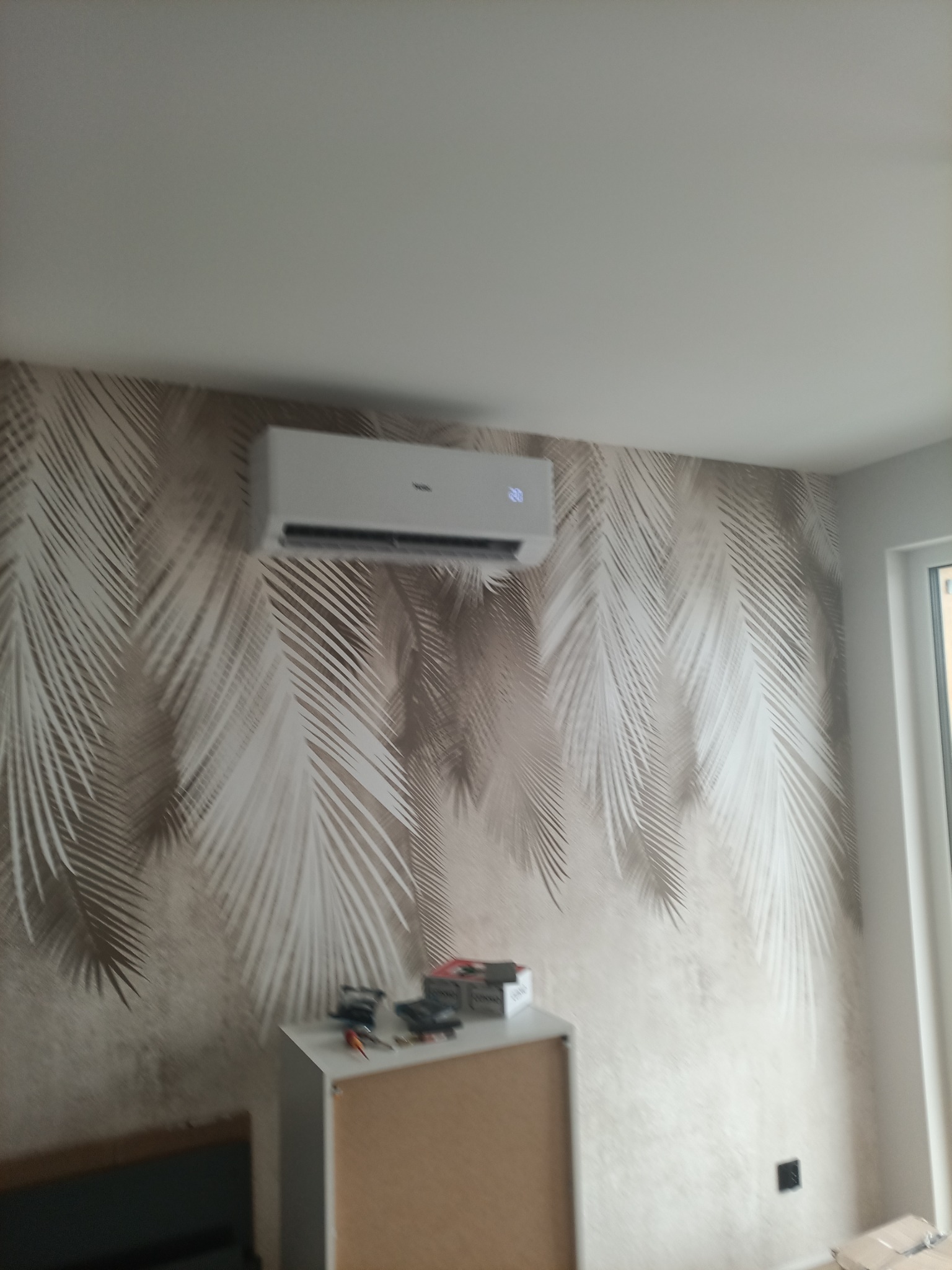 Zamontowany klimatyzator na ścianie z tapetą w palmy. Na szafce pod nim narzędzia. Widoczny fragment okna i gniazdko elektryczne. Ujęcie z dołu.