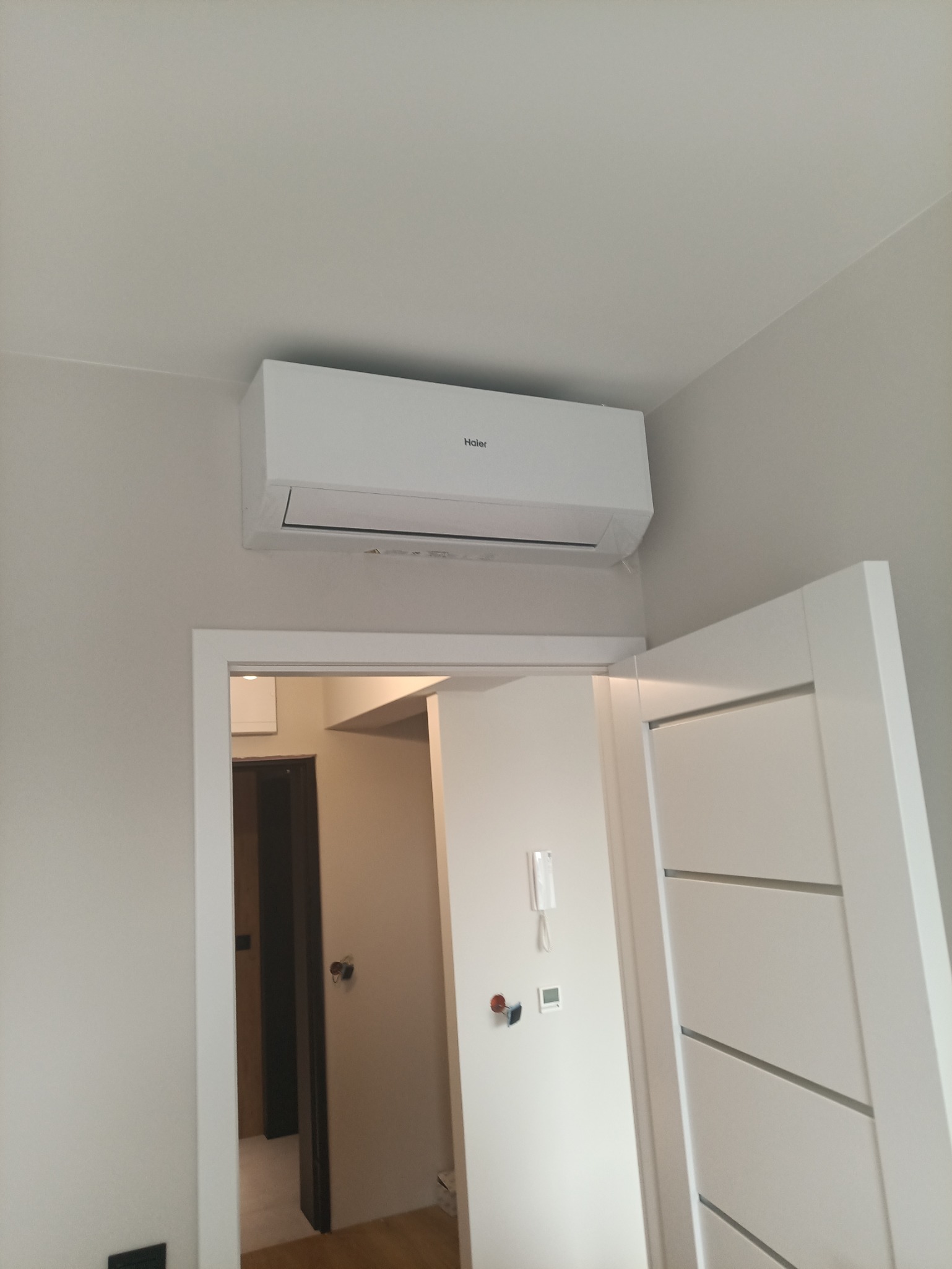 Biała klimatyzacja Haier zamontowana nad drzwiami w jasnym wnętrzu. Widoczny panel sterowania na ścianie i fragment otwartych drzwi.