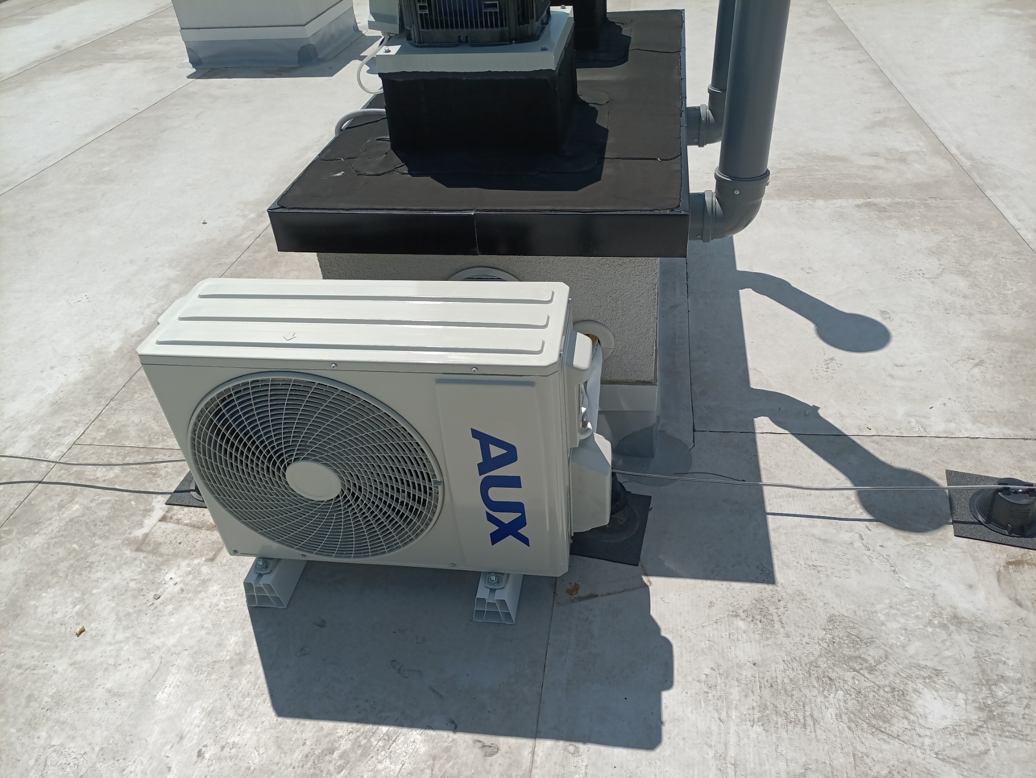 Biała jednostka zewnętrzna klimatyzacji AUX na płaskim dachu, widoczne elementy instalacji i cień. Jasne, naturalne światło.