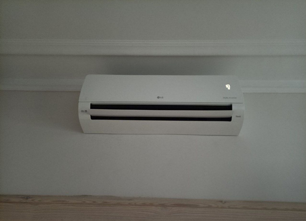 Biała klimatyzacja LG, zamontowana na ścianie w jasnym pomieszczeniu. Widoczny wyświetlacz temperatury i logo producenta. Minimalistyczny design.