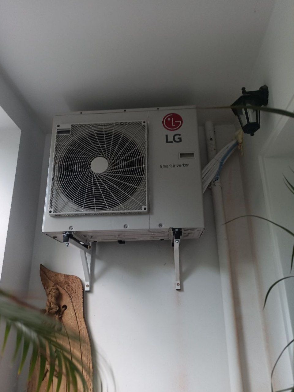 Biała jednostka zewnętrzna klimatyzacji LG Smart Inverter zamontowana na ścianie, widoczne kable i lampa ścienna. Dekoracja z drewna w tle.
