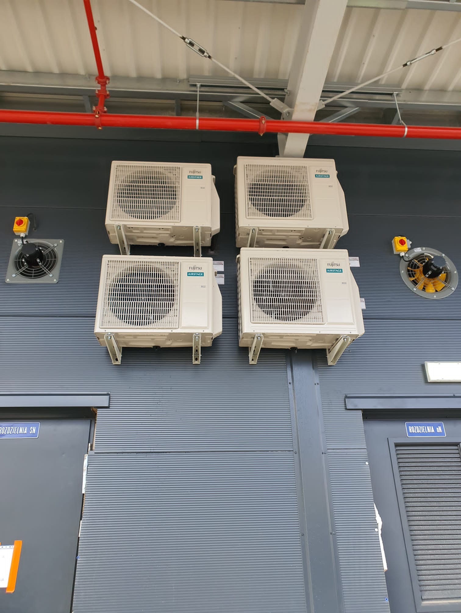 Cztery klimatyzatory Fujitsu Airstage zamontowane na szarej, blaszanej ścianie budynku przemysłowego, z widocznymi wentylatorami i czerwoną rurą instalacyjną.