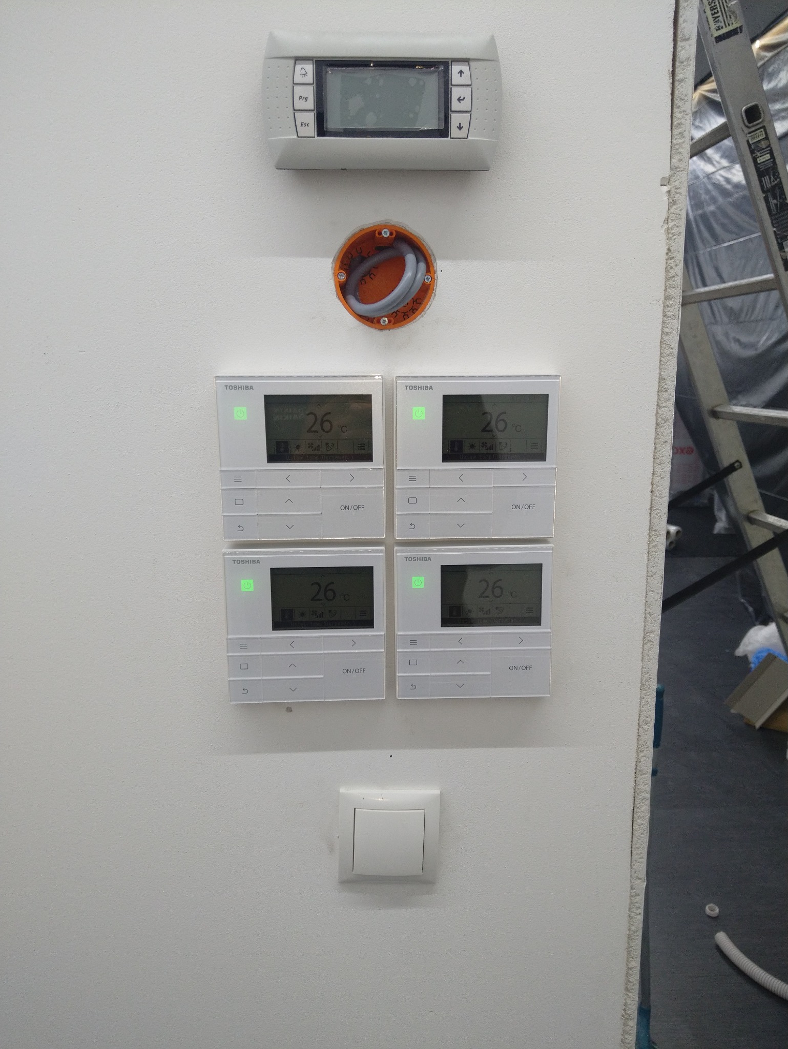 Cztery panele sterowania klimatyzacją Toshiba zamontowane na białej ścianie, nad nimi panel główny i puszka instalacyjna. Widoczna drabina w tle.