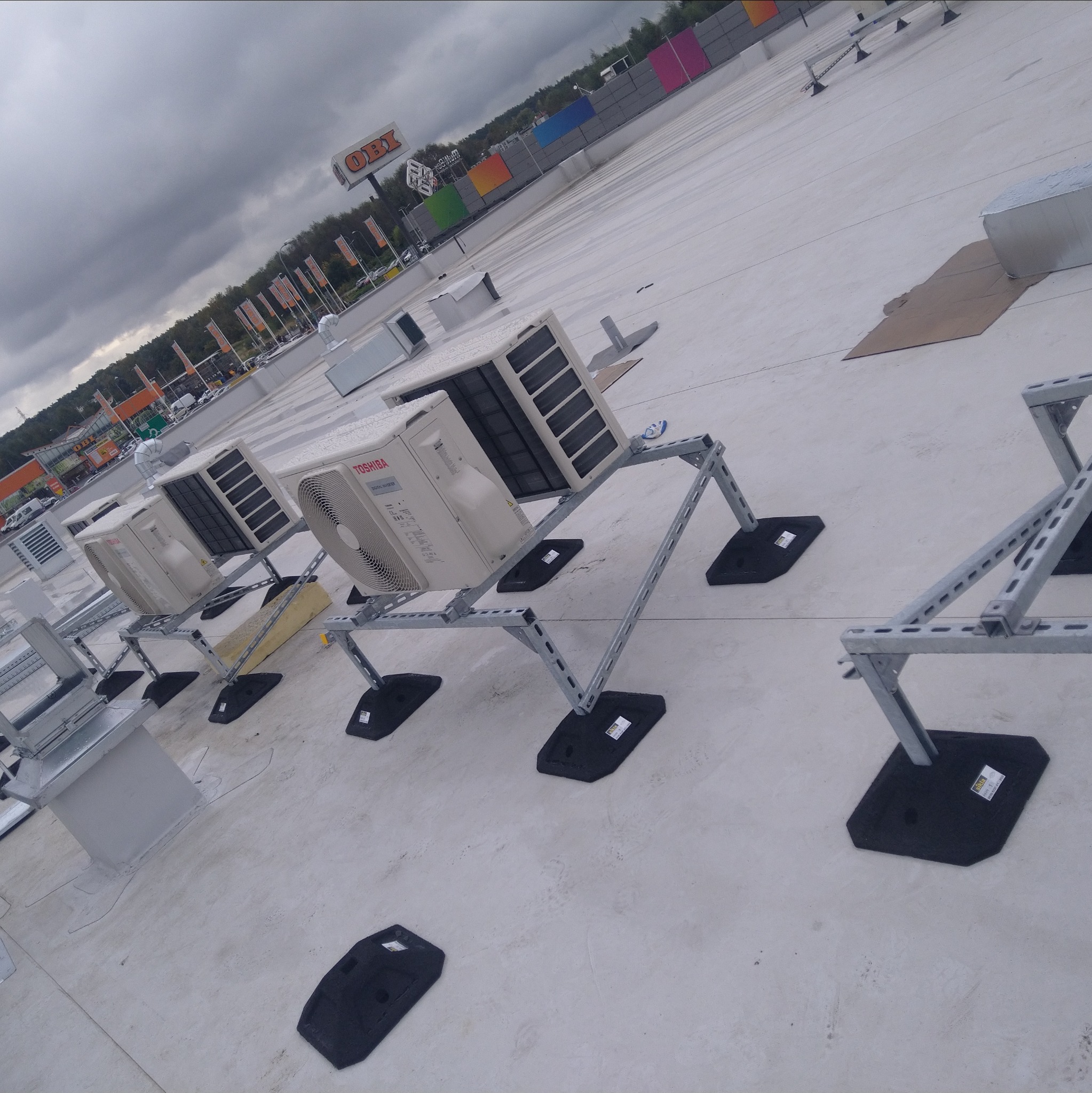 Instalacja klimatyzacji Toshiba na dachu budynku z widokiem na sklep OBI w pochmurny dzień. Metalowe konstrukcje wsporcze i czarne podkładki antywibracyjne.