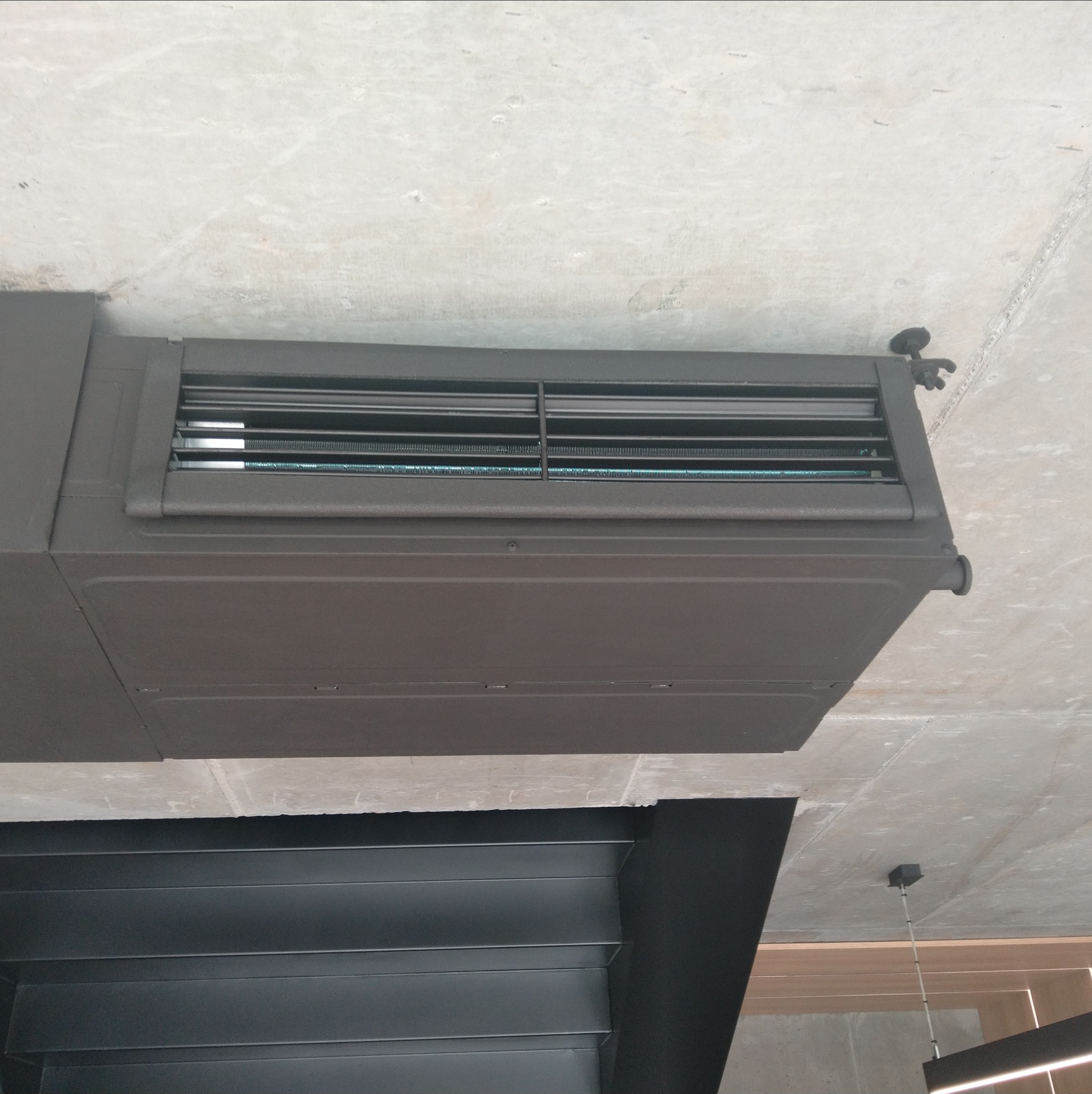 Zamontowana, brązowa klimatyzacja kanałowa na betonowym suficie. Widoczne kratki wentylacyjne i elementy mocujące. Ujęcie z dołu, w tle fragment wnętrza.