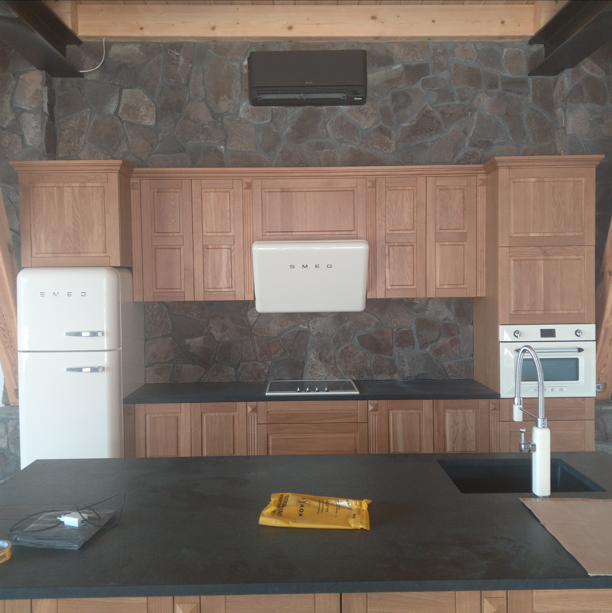Kuchnia w stylu retro z drewnianymi szafkami, sprzętem SMEG i kamienną ścianą. Widoczny klimatyzator Daikin nad szafkami. Na blacie leży folia ochronna.