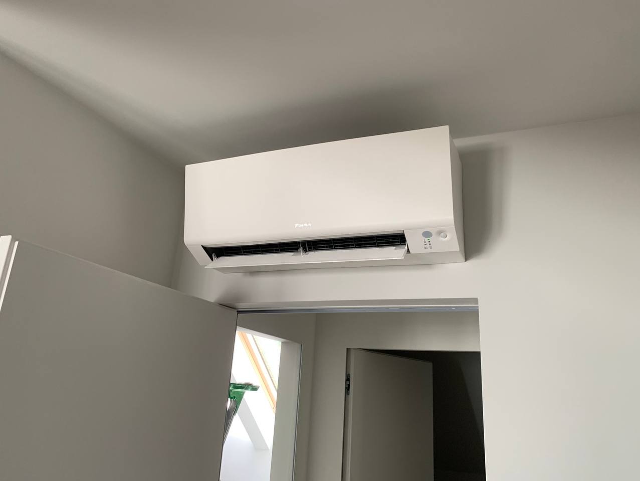 Biała klimatyzacja Daikin zamontowana na ścianie nad otwartymi drzwiami. Widoczny fragment poddasza przez drzwi. Minimalistyczny design, jasne wnętrze.