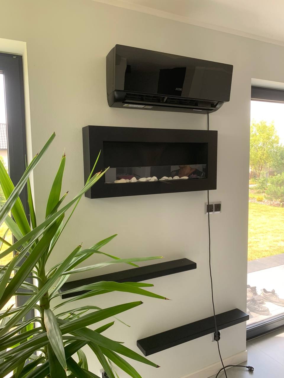 Nowoczesny, czarny klimatyzator ścienny marki AUX, zamontowany nad elektrycznym kominkiem w salonie z minimalistycznym wystrojem i zieloną rośliną doniczkową.