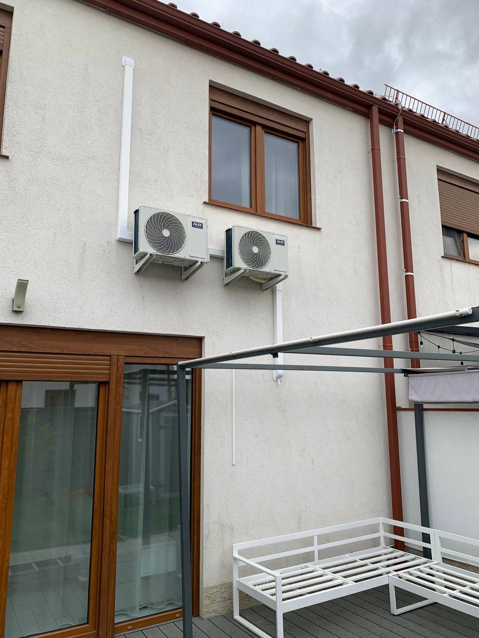 Dwa klimatyzatory AUX zamontowane na elewacji domu z brązowymi oknami i rynnami. Białe rury odprowadzające skropliny. Widoczny fragment tarasu z pergolą i białą ławką.