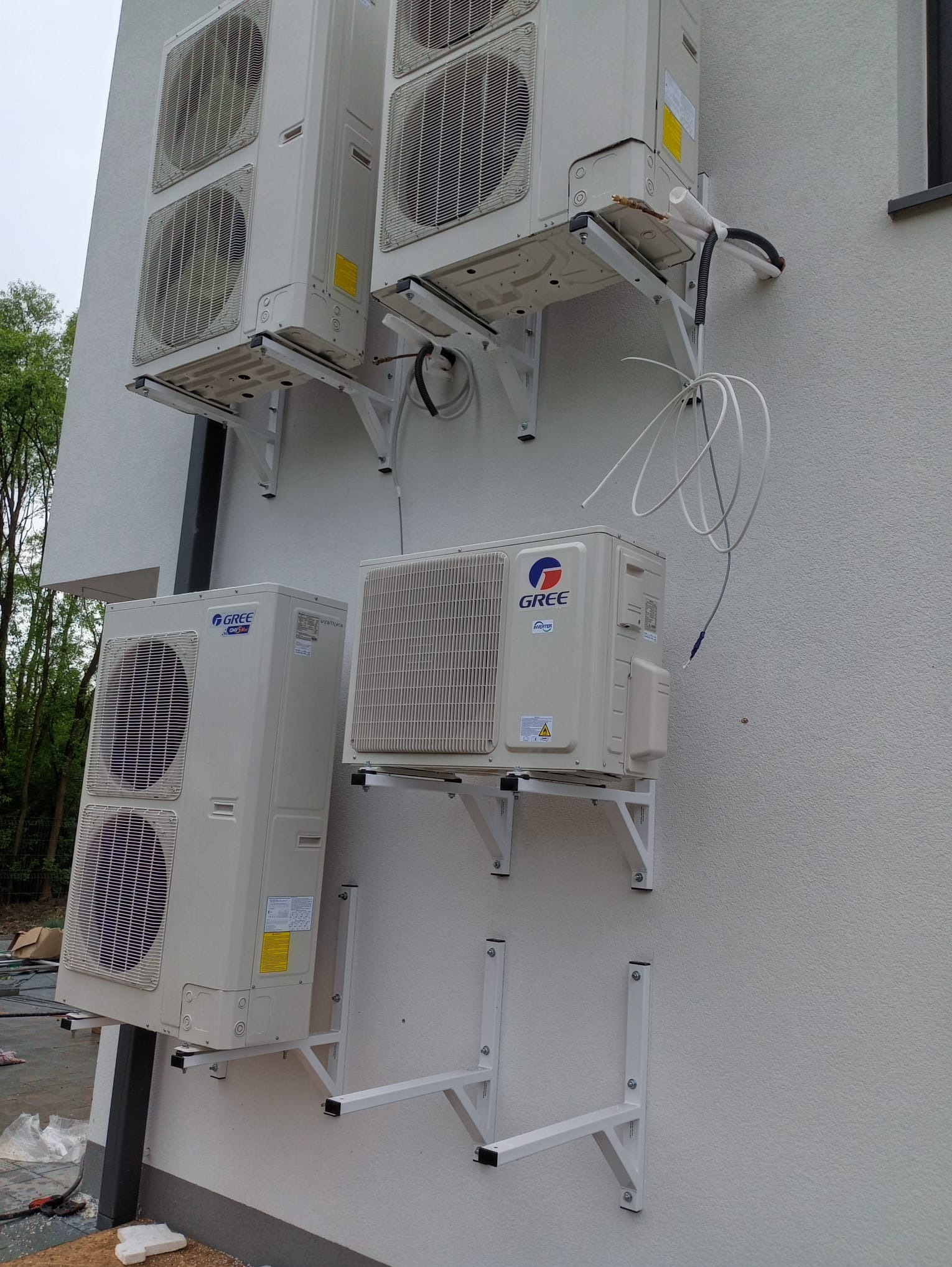 Cztery klimatyzatory marki Gree zamontowane na białej ścianie budynku, widoczne wsporniki i instalacje. Ujęcie z dołu, częściowo widoczne drzewa w tle.