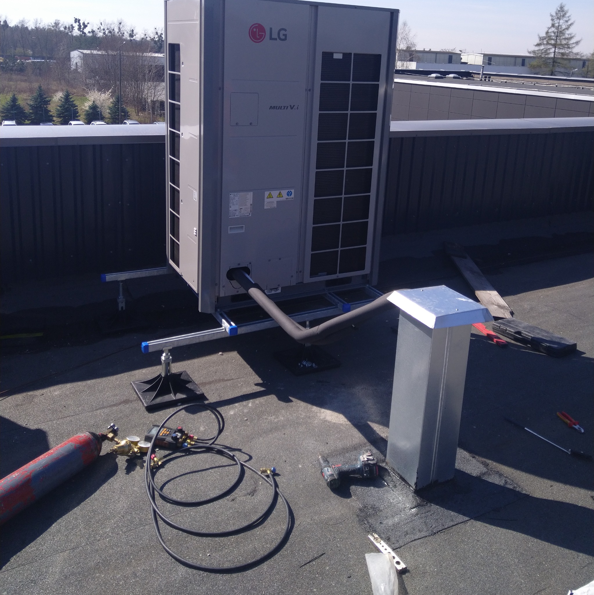 Montaż zewnętrznej jednostki klimatyzacji LG Multi V i na dachu budynku. Widoczne narzędzia i elementy instalacji, w tle zabudowa miejska i zieleń.
