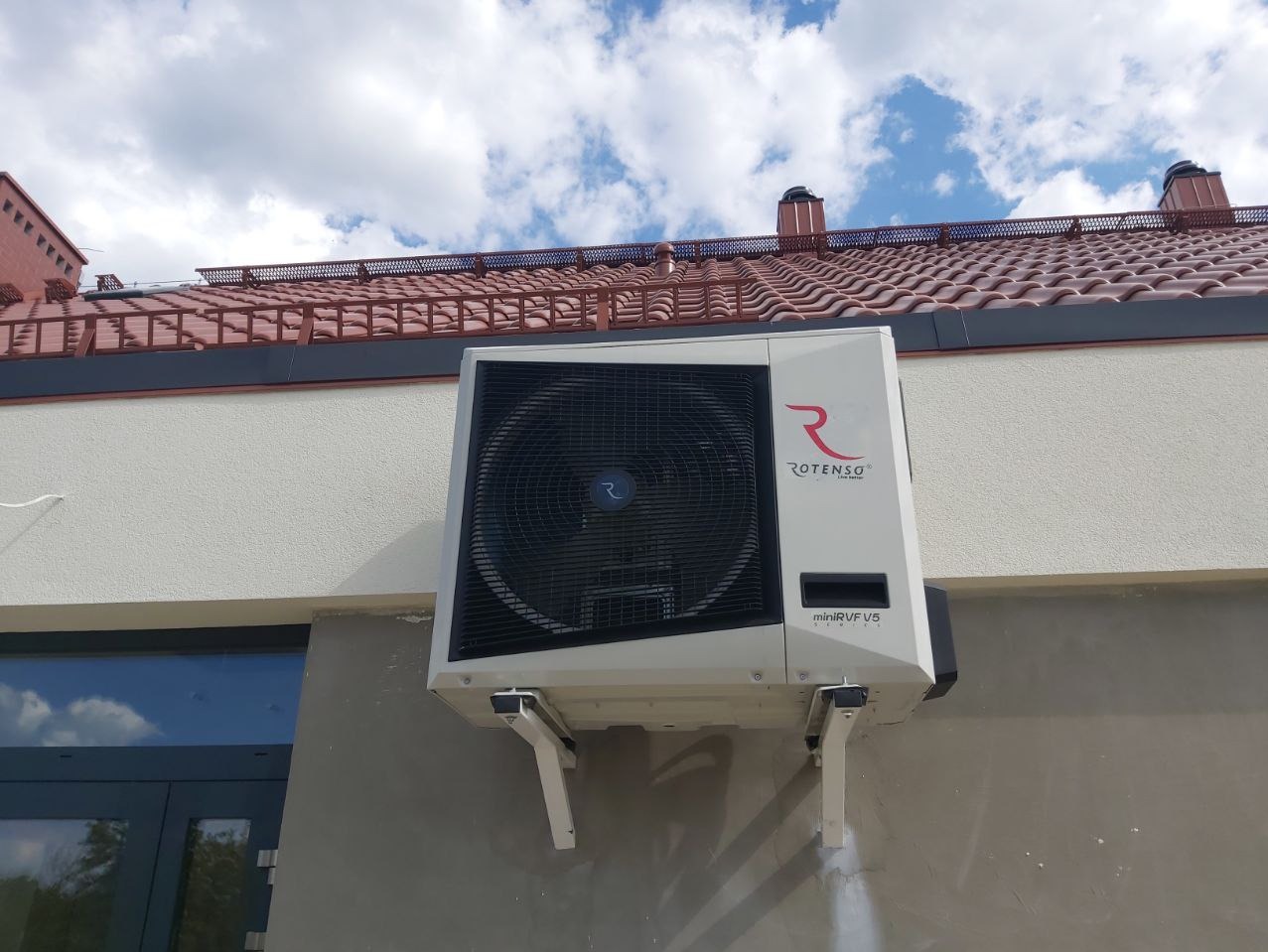 Biała jednostka zewnętrzna klimatyzacji Rotenso miniRVR V5 zamontowana na ścianie budynku z czerwoną dachówką, widoczna na tle błękitnego nieba z chmurami.