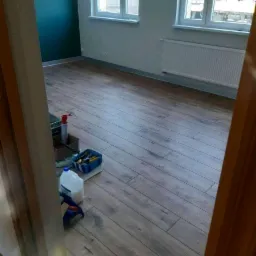 Pokój po ułożeniu paneli podłogowych z widocznymi narzędziami i materiałami budowlanymi w rogu, ściana pomalowana na ciemny zielony kolor, dwa okna z widokiem na zewnątrz.