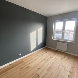 Cezary Matuszewski - Pusty pokój po remoncie: ściana pomalowana na ciemny grafit, jasne panele podłogowe z widoczną strukturą drewna, białe listwy przypodłogowe, okno z widokiem na budynki, grzejnik pod oknem, gniazdko...
