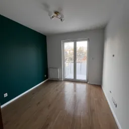 Pusty pokój po remoncie z drewnianą podłogą, ścianą pomalowaną na ciemnozielony kolor i białymi ścianami oraz oknem balkonowym.