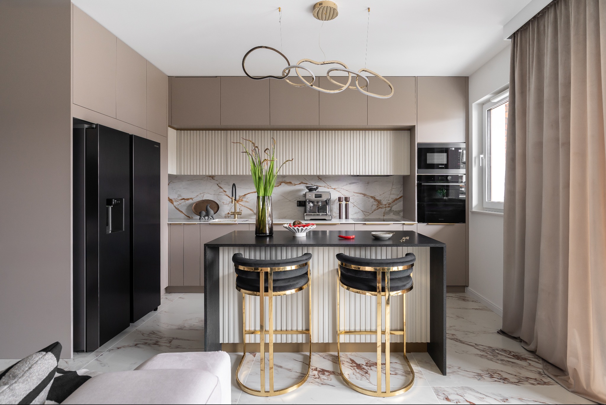 Elegancka, nowoczesna kuchnia z marmurowym blatem, czarną wyspą i złotymi hokerami. Beżowe szafki i oryginalna lampa wisząca tworzą spójną całość.