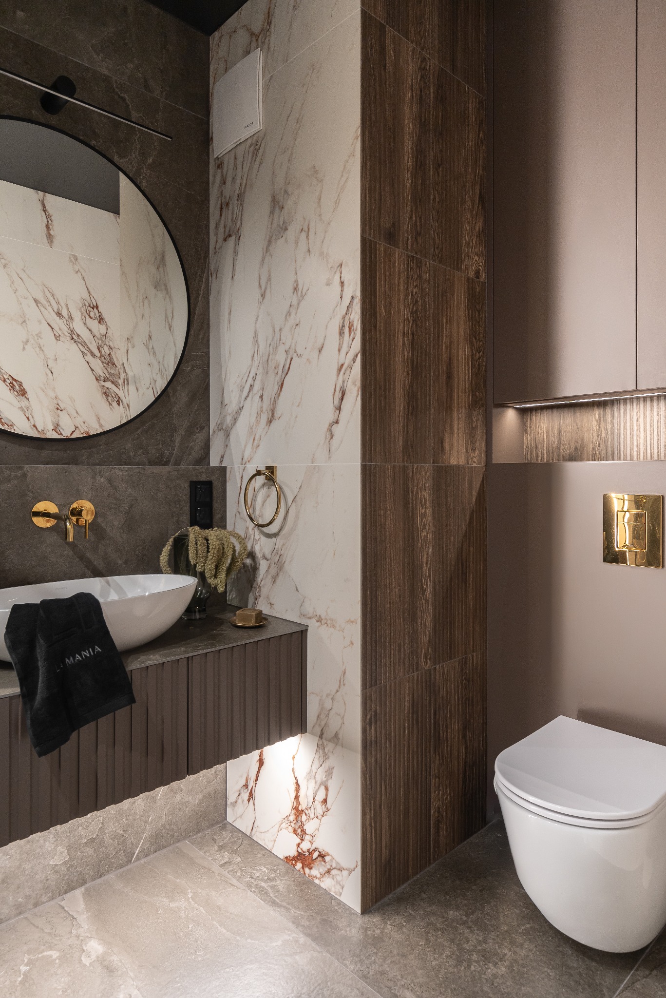 Elegancka łazienka z marmurowymi płytkami, okrągłym lustrem i złotymi akcentami. Umywalka nablatowa, toaleta podwieszana, oświetlenie LED. Nowoczesny design.