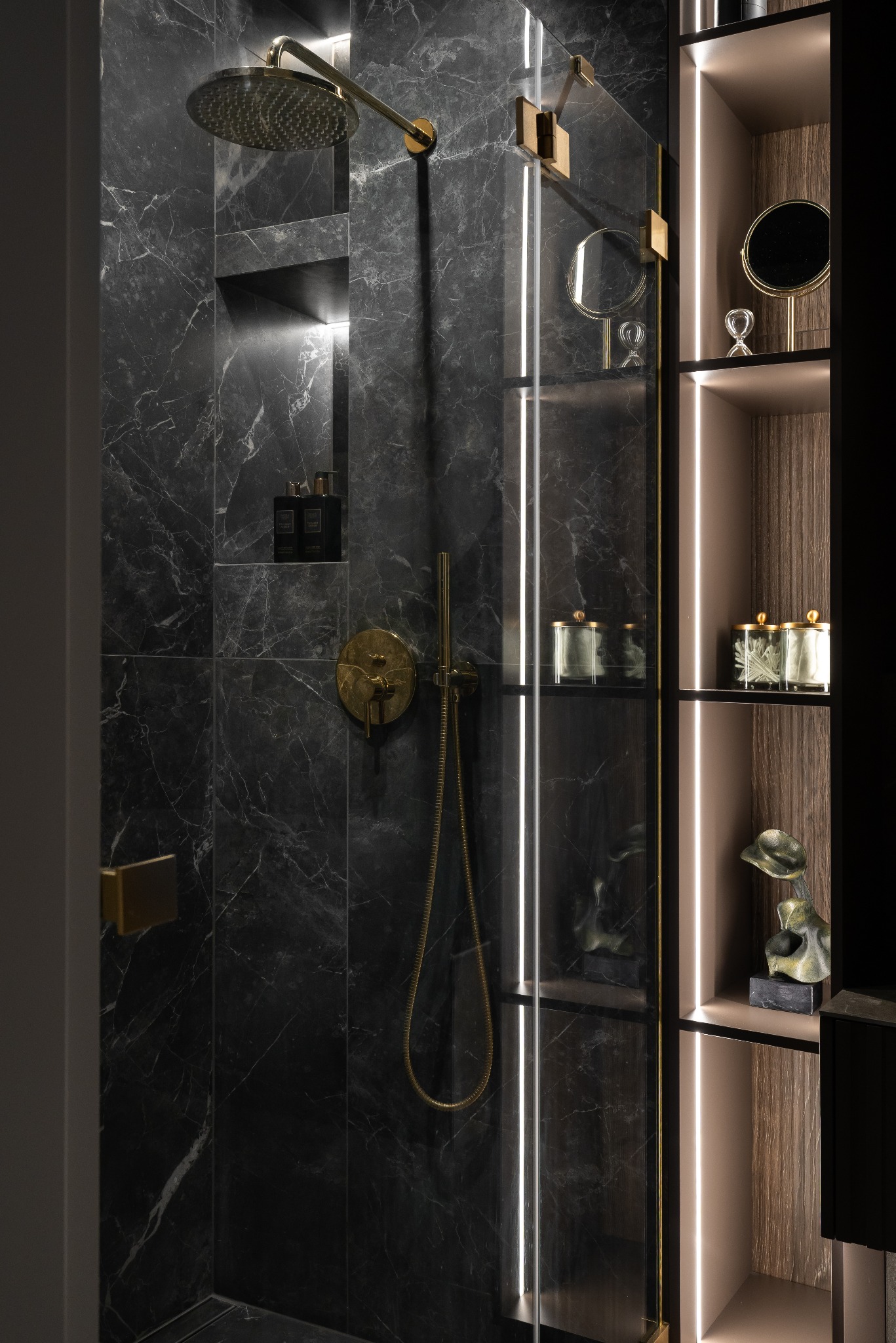 Elegancka kabina prysznicowa z czarnych marmurowych płytek, złota armatura, oświetlone półki z dekoracjami. Nowoczesny design łazienki, luksusowe wykończenie.