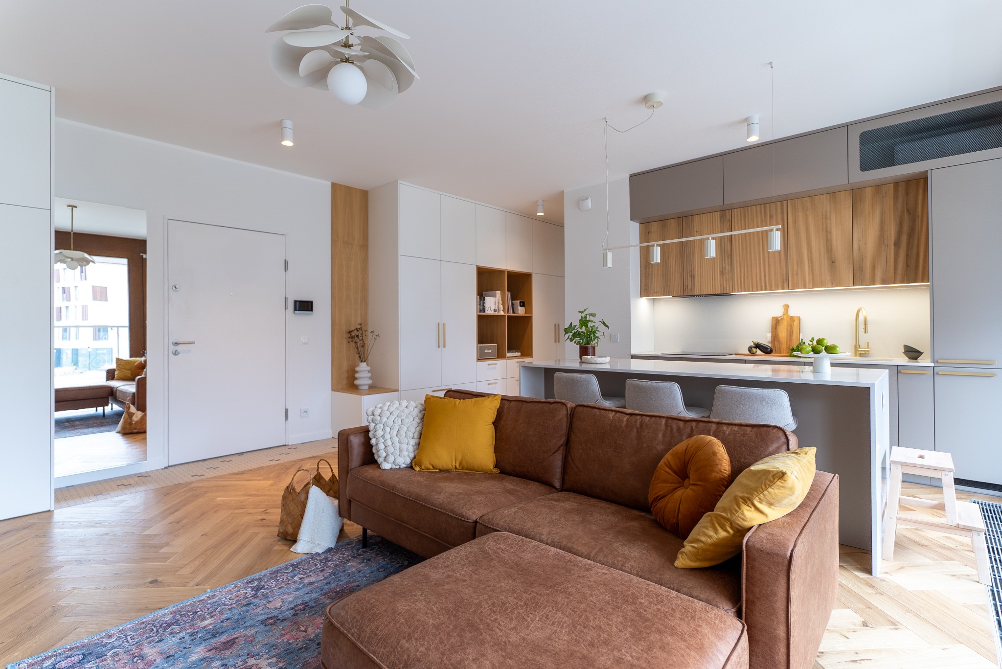 Stylowe wnętrze: brązowa sofa z poduszkami, drewniana podłoga w jodełkę, biała zabudowa kuchenna z drewnianymi akcentami i wyspą. Nowoczesne oświetlenie.