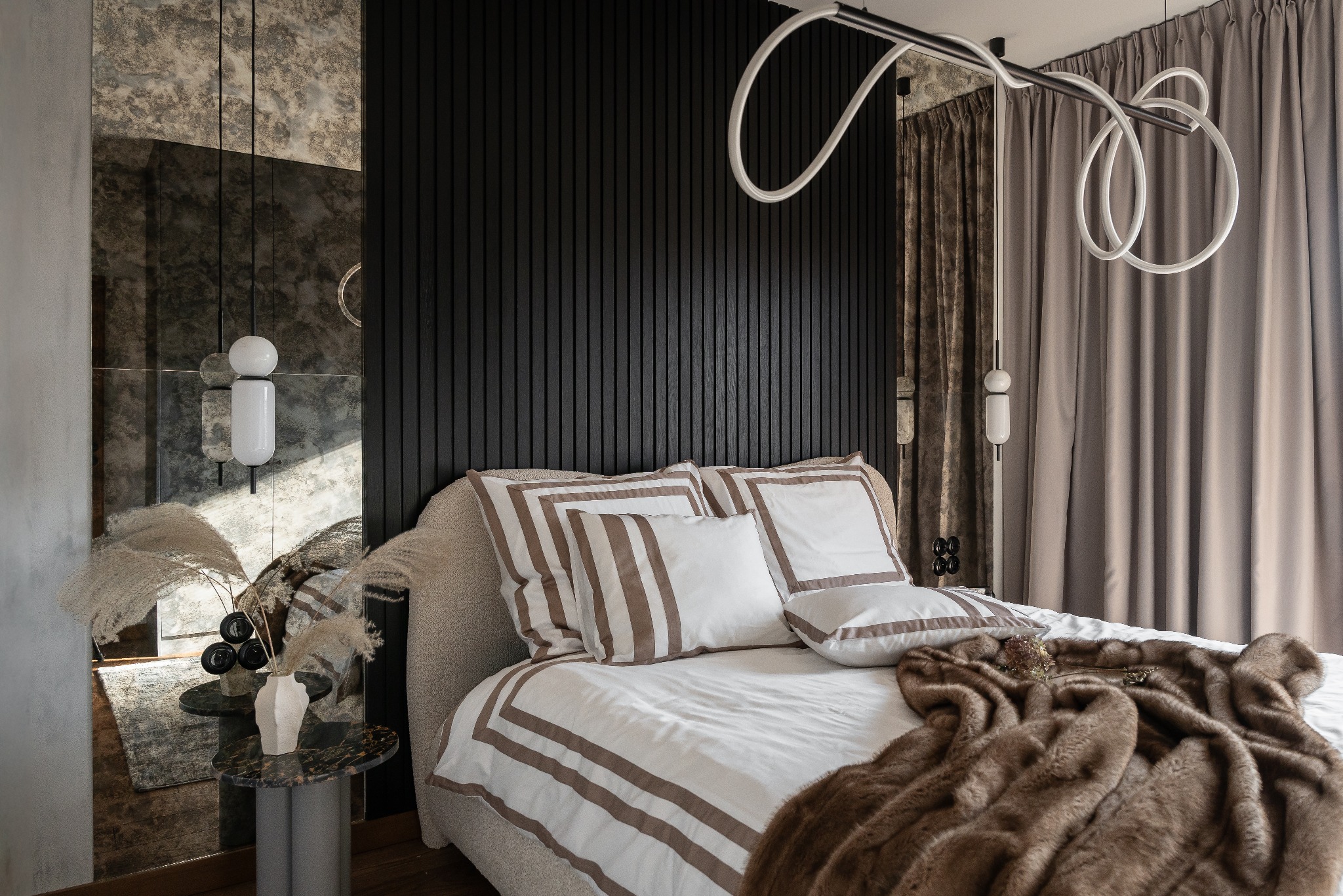 Stylowa sypialnia z łóżkiem, czarną ścianą z pionowych desek i lustrem z efektem patyny. Dekoracyjne oświetlenie i zasłony dodają elegancji.