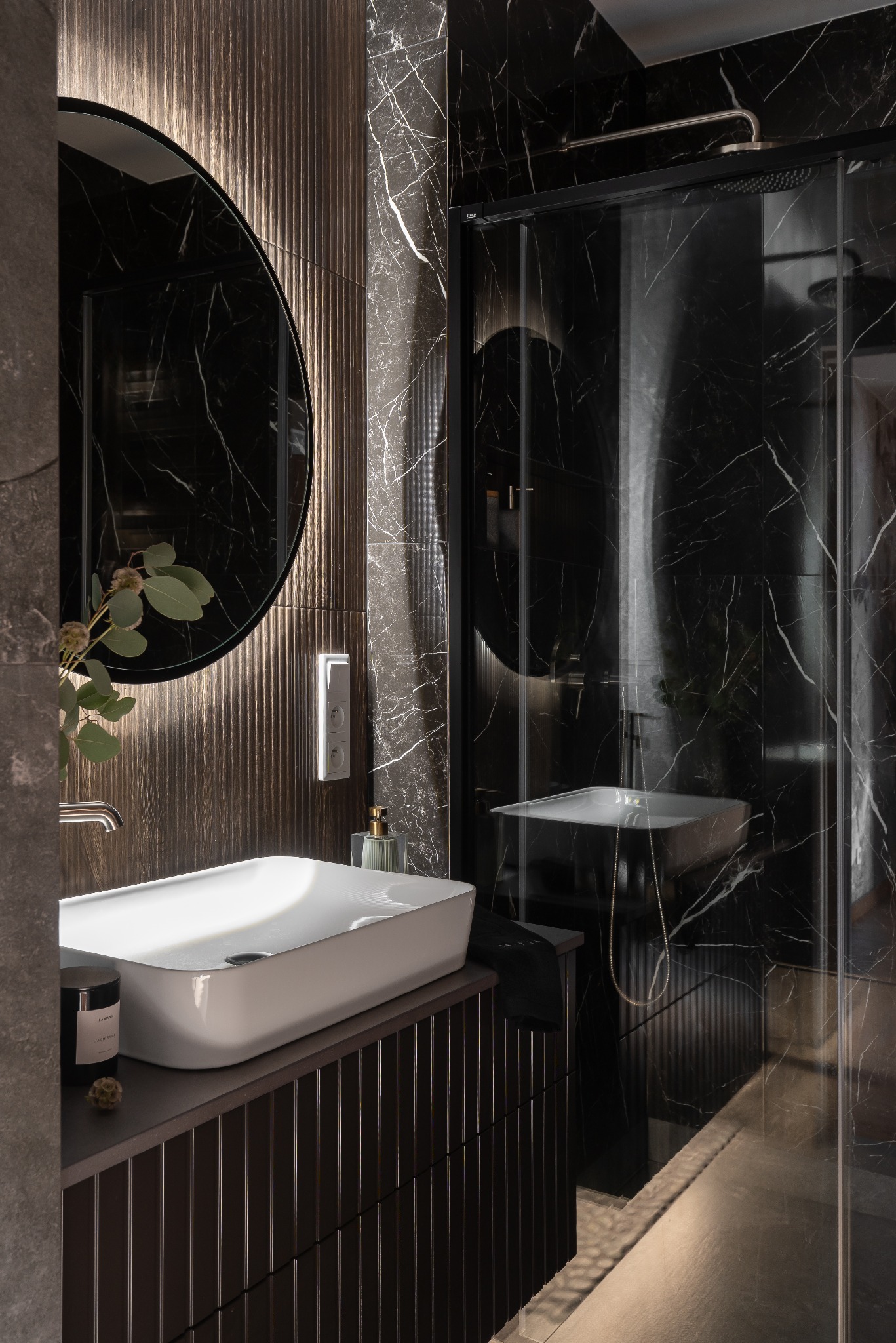 Ekskluzywna łazienka z czarnym marmurem, owalnym lustrem z podświetleniem i białą umywalką nablatową. Nowoczesny design z akcentami złota i roślinnym detalem.