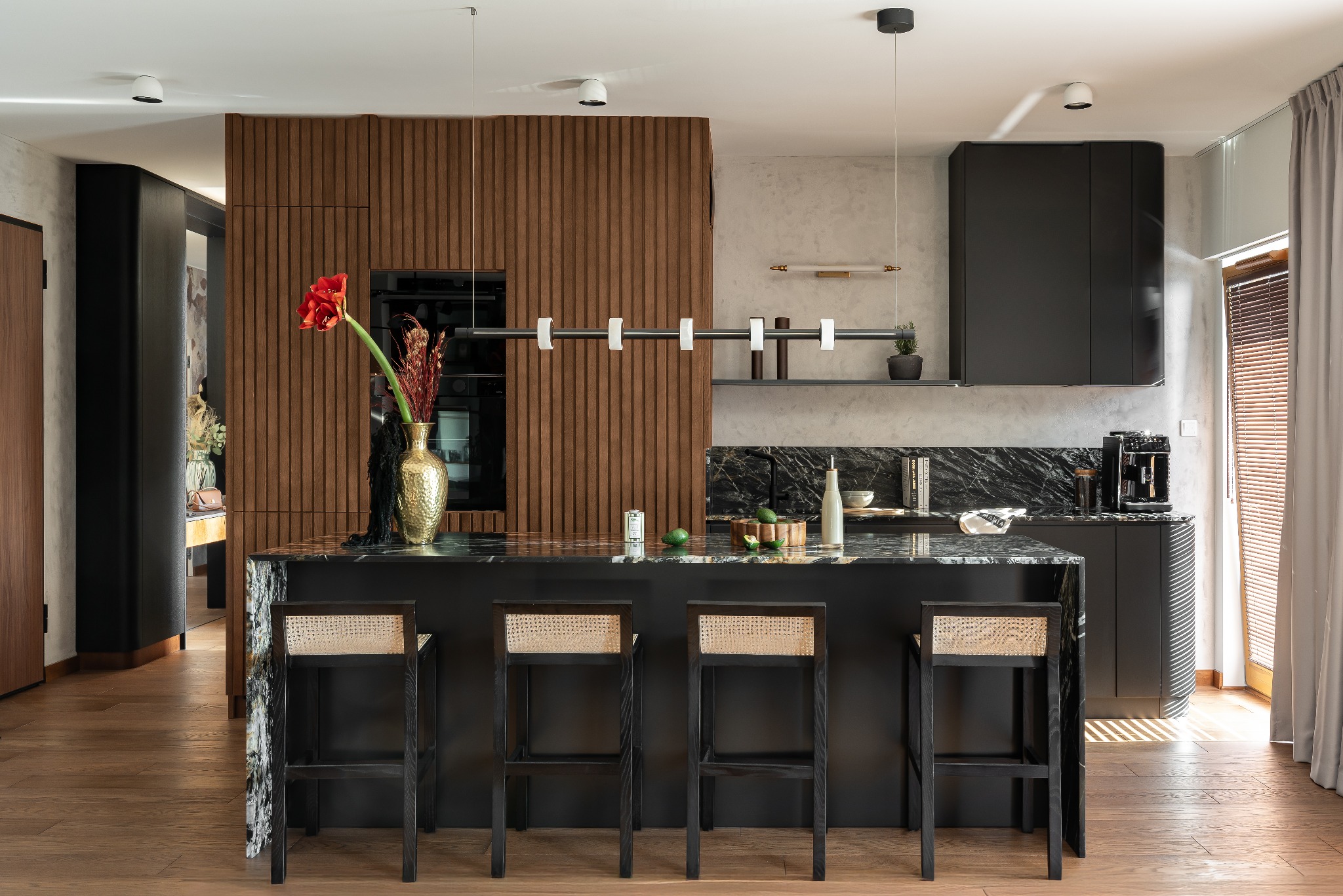 Nowoczesna kuchnia z wyspą z czarnego marmuru i hokerami z rattanu. Drewniana ściana z pionowymi panelami i oświetlenie liniowe. Minimalistyczny design wnętrza.