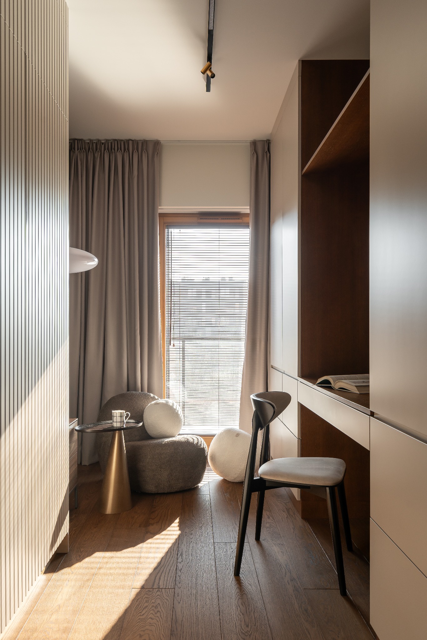 Nowoczesny pokój z drewnianą podłogą, biurkiem z otwartą książką, krzesłem i pufami przy oknie z żaluzjami. Ściana z panelami i designerska lampa dodają charakteru.