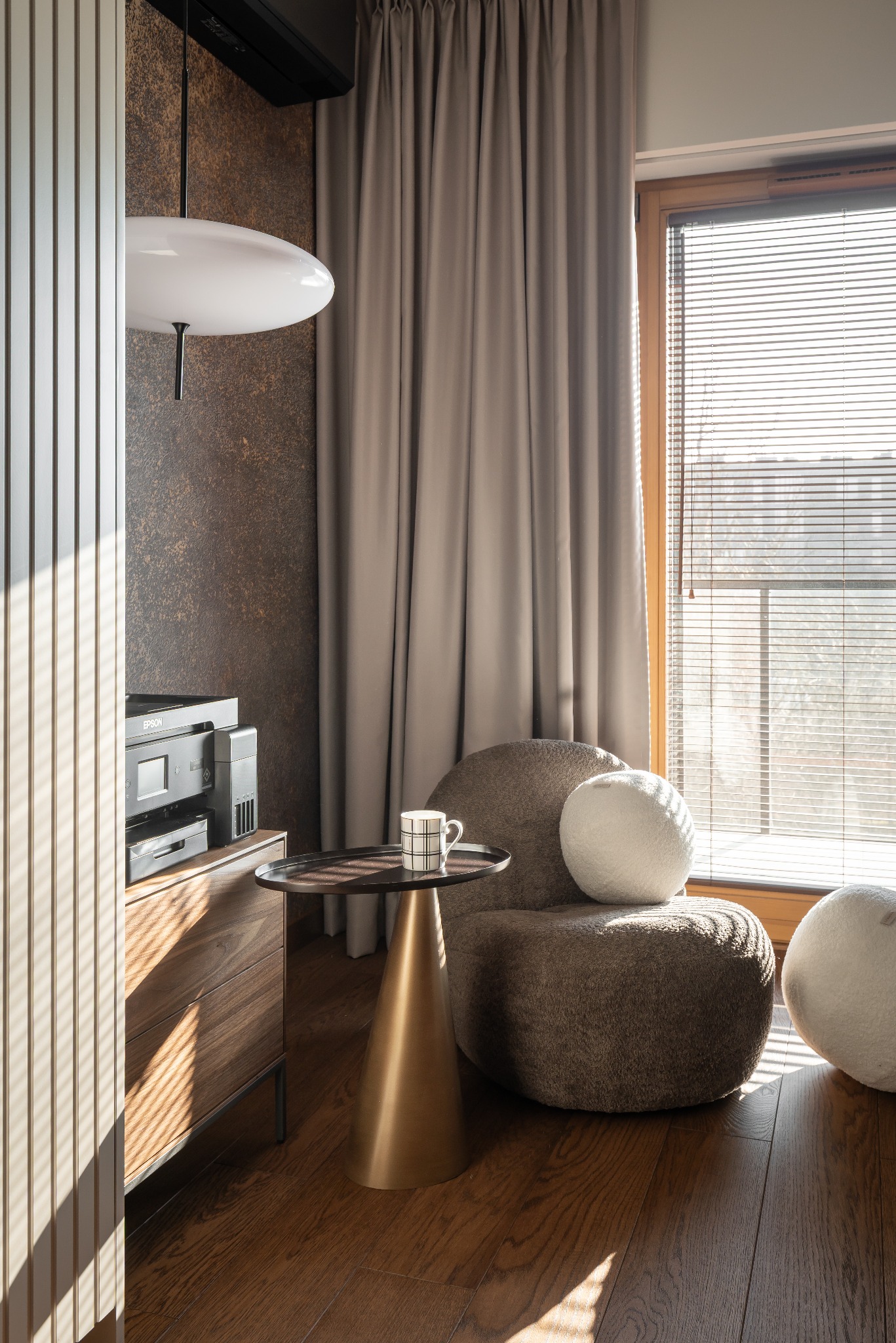 Stylowe wnętrze z brązową ścianą, drewnianą podłogą i designerskim fotelem. Uwagę przykuwa złoty stolik z filiżanką. Okno z żaluzjami wpuszcza jasne światło.